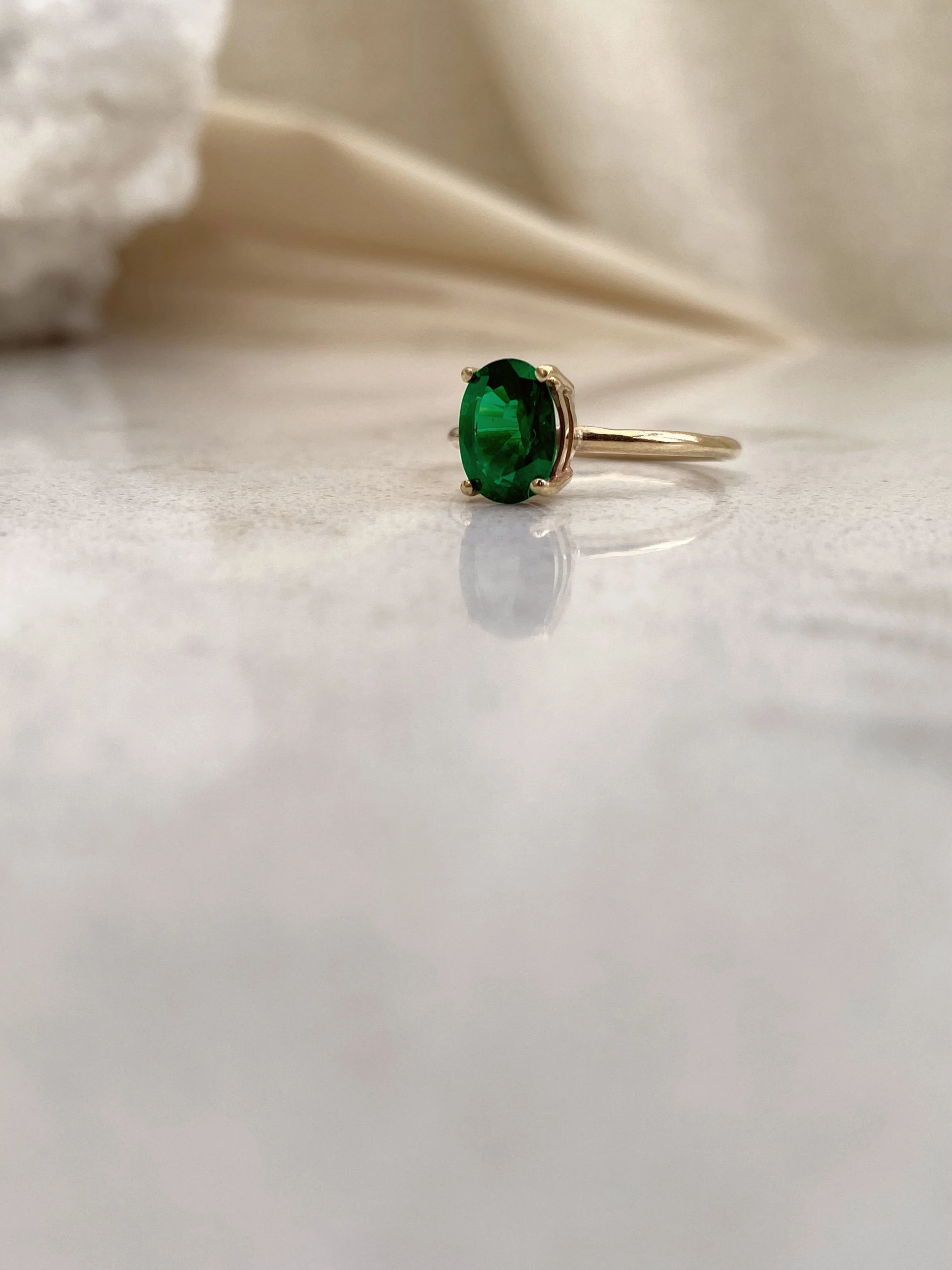 A gold ring by Amy Russell Taylor with a large oval green gemstone, placed on a smooth surface with a blurred, light-colored background.