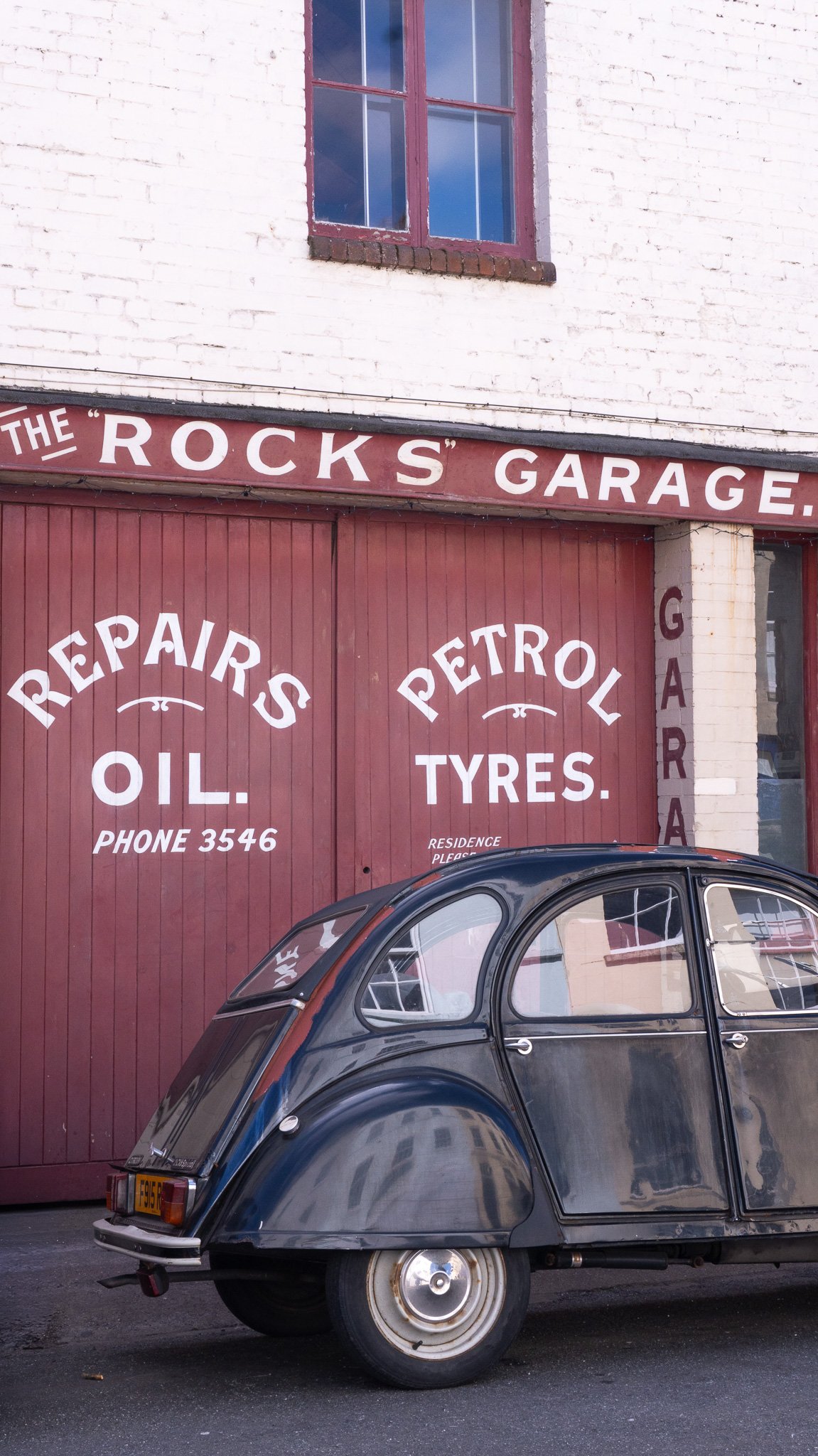 A vintage black car parked in front of a garage with red doors. The garage has white painted brick walls, and signs advertising oil, tires, and repairs. The signage says The Rocks Garage in Clifton, Bristol