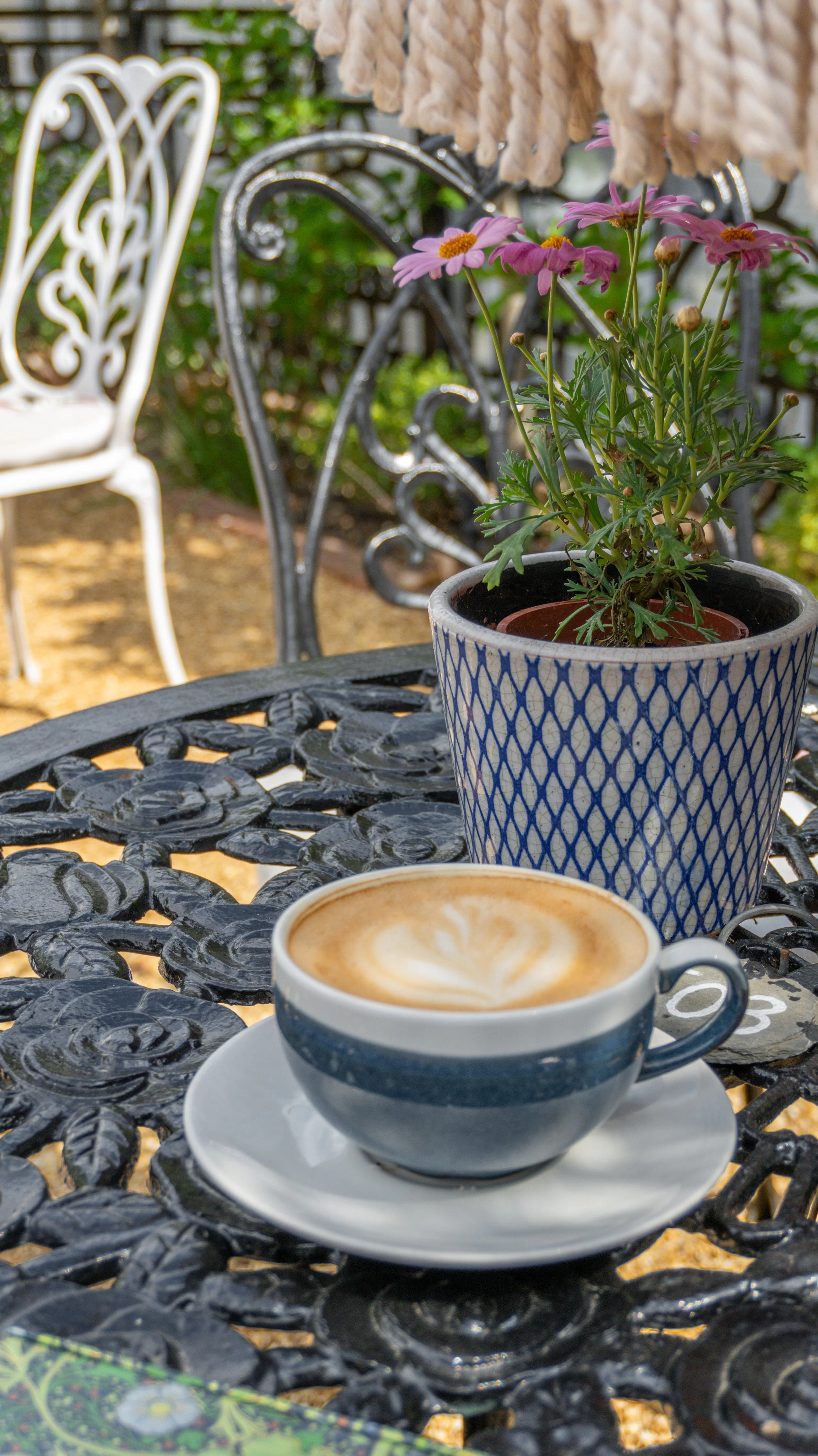 A table at Stanwell House with a cup of coffee with latte art, a blue patterned coffee mug with pink flowers, and a potted plant with pink flowers on an outdoor black wrought iron table and chairs.