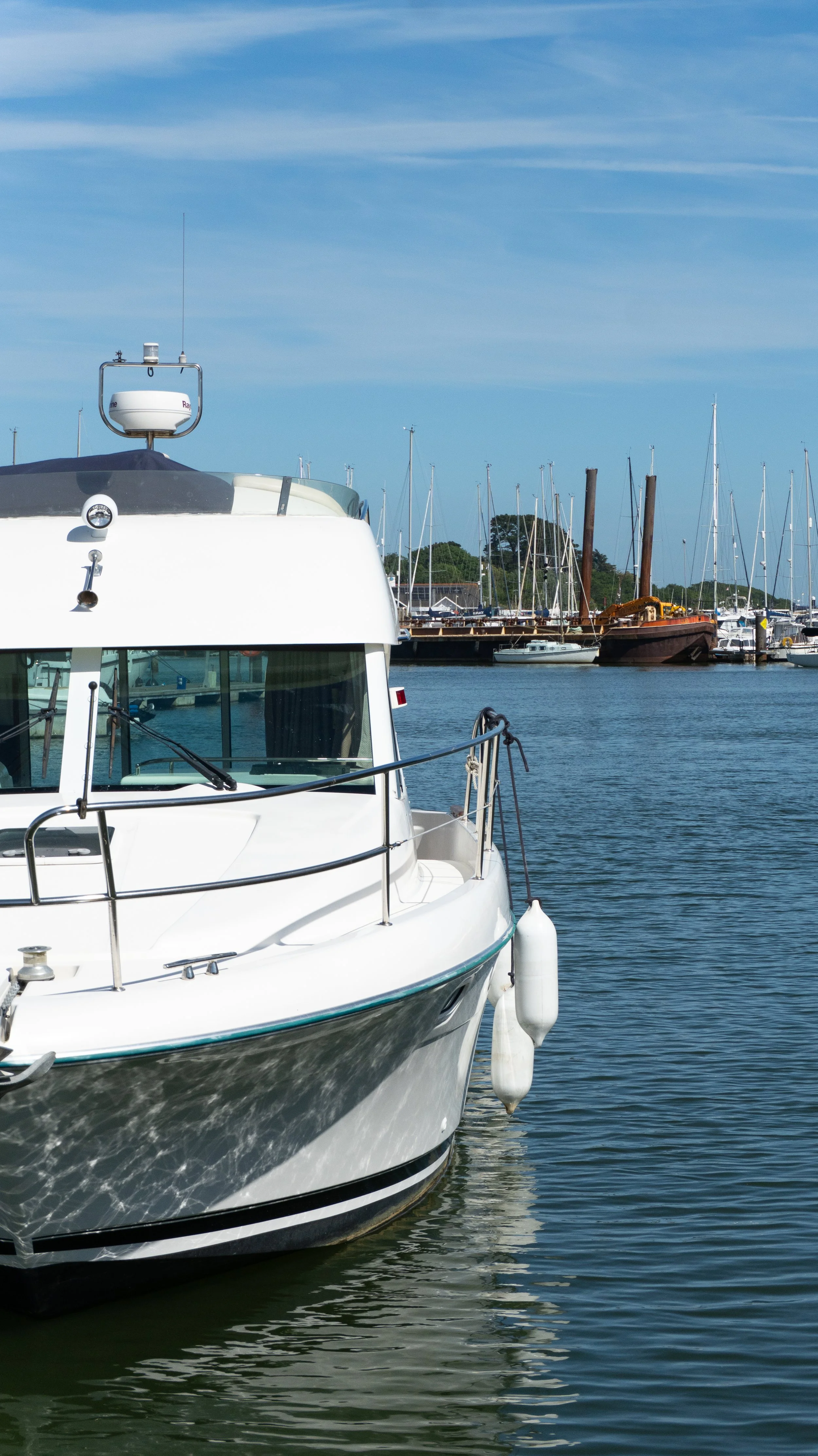 Close-up of a white boat docked at Lymington marina with other sailboats and boats in the background under a partly cloudy sky.
