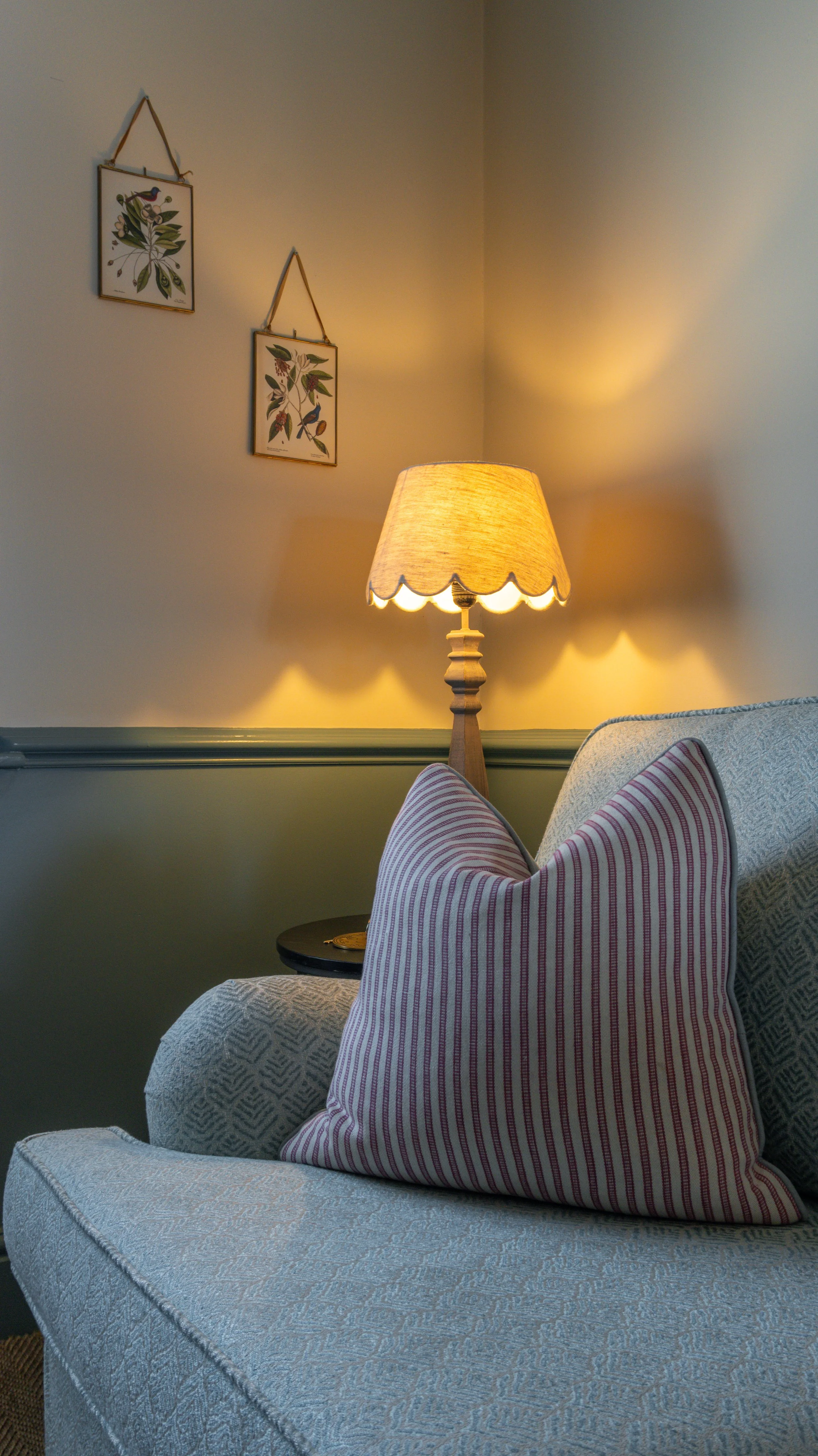 A cozy room corner at Stanwell House with a beige armchair, a pink and white striped pillow, a side table, a lamp with a beige shade, and framed botanical art on a light yellow wall.
