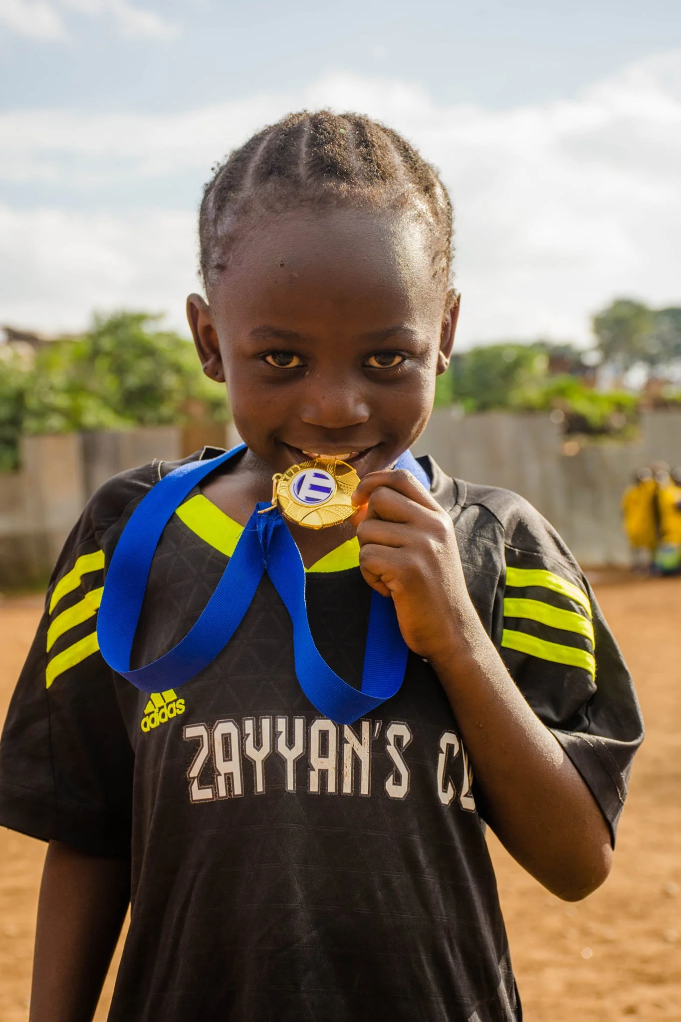 A young student from KIMMTA holding a gold medal with a blue ribbon, smiling slightly, standing outdoors on a dirt field with trees and buildings in the background.