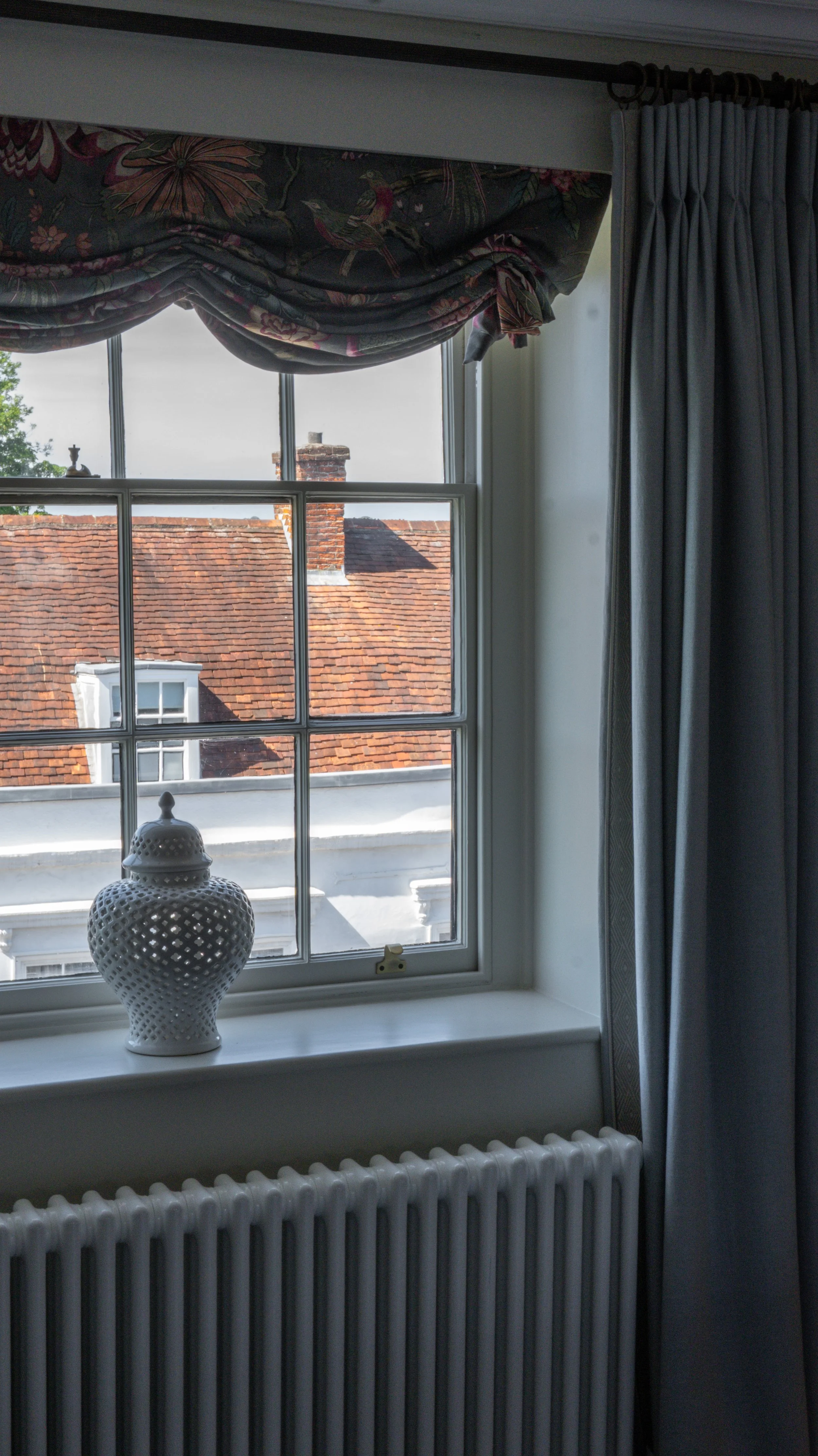 Interior view of a room window at Stanwell House with a floral curtain valance and side curtains, a small decorative ceramic lamp on the windowsill, and a view of rooftops with a brick chimney outside.