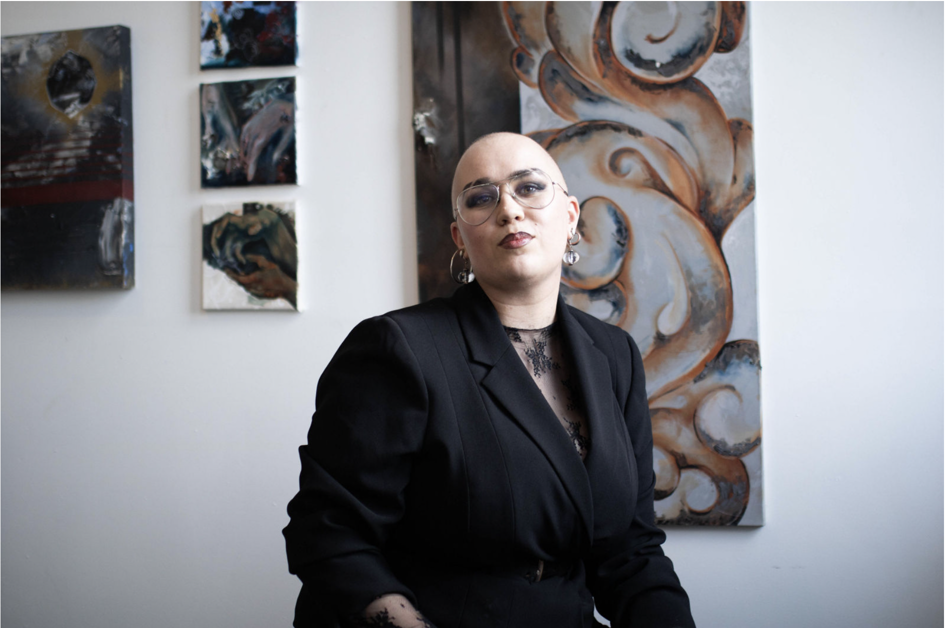A person with a shaved head and glasses wearing a black blazer and lace top, sitting in front of abstract artwork on a white wall.