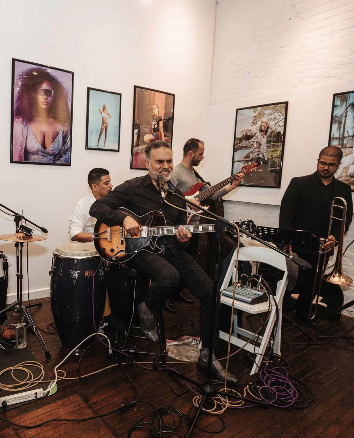 A band performs live music in an art gallery. The lead singer is playing an acoustic guitar, with other band members playing percussion, electric guitar, and trombone. The gallery walls are decorated with framed photographs of fashion and travel.