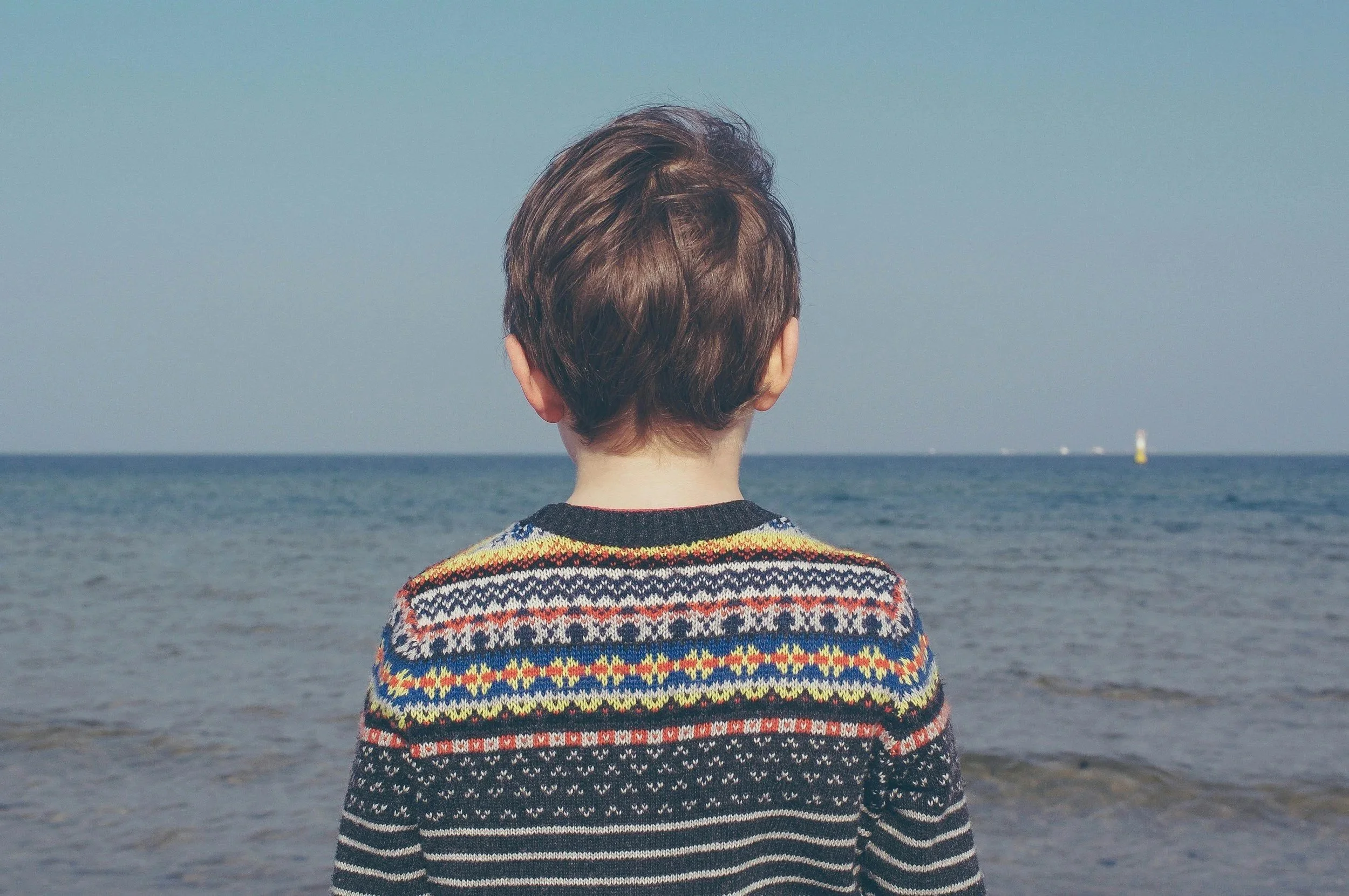 A young person with short brown hair faces away from the camera and is wearing a black knit sweater with brightly colored stripes. They are looking out over the ocean.