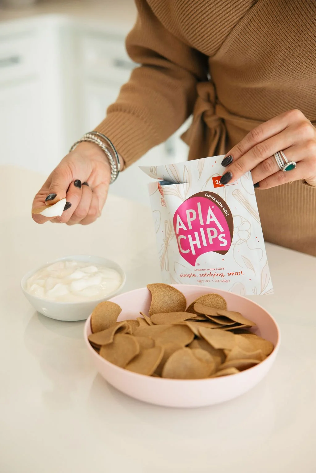 Person opening a bag of Apia Chips cinnamon roll flavored almond flour chips, with a pink bowl of chips and a small bowl of yogurt on a white table.