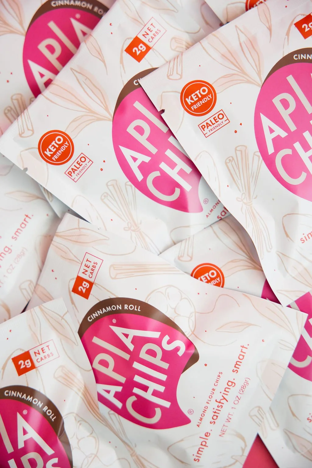 Multiple packets of APIA Chips cinnamon roll flavored almond flour chips with pink and white packaging, featuring labels indicating 2 grams of net carbs and keto-friendly and paleo-friendly attributes.
