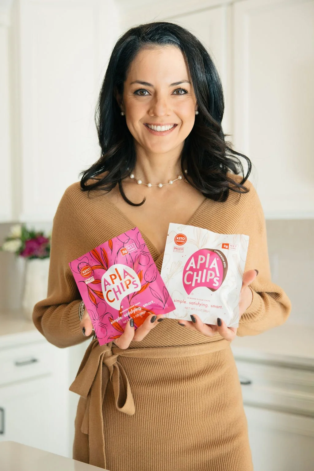 Woman smiling and holding two bags of APIA Chips, one pink and one white, in a kitchen setting.