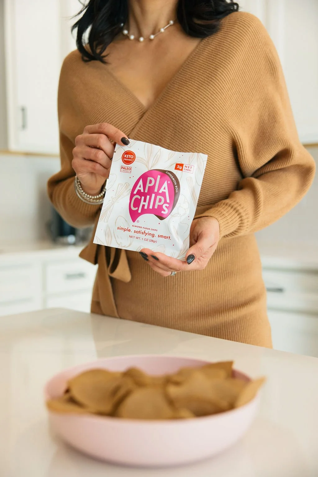 A woman holding a bag of Apia Chips cinnamon roll flavor almond flour chips in a kitchen, with a pink bowl of chips on the countertop.