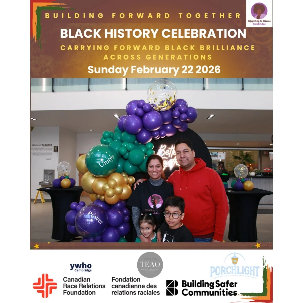 Family of four smiling at a black table surrounded by colorful balloons with words like 'Unity' and 'Power,' at a Black History Celebration event on February 22, 2026, with a sign in the background reading 'Building Forward Together' and various orga