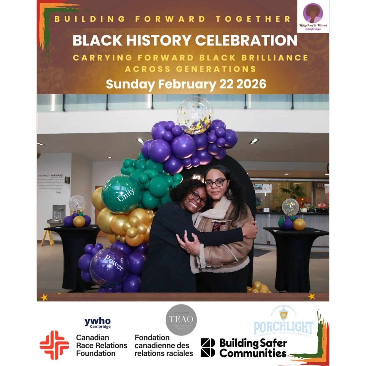 Two women hugging in front of a display of purple, green, gold, and clear balloons with a large sign for a Black History Celebration event on February 22, 2026, inside a building. The sign includes text about building forward together and carrying fo