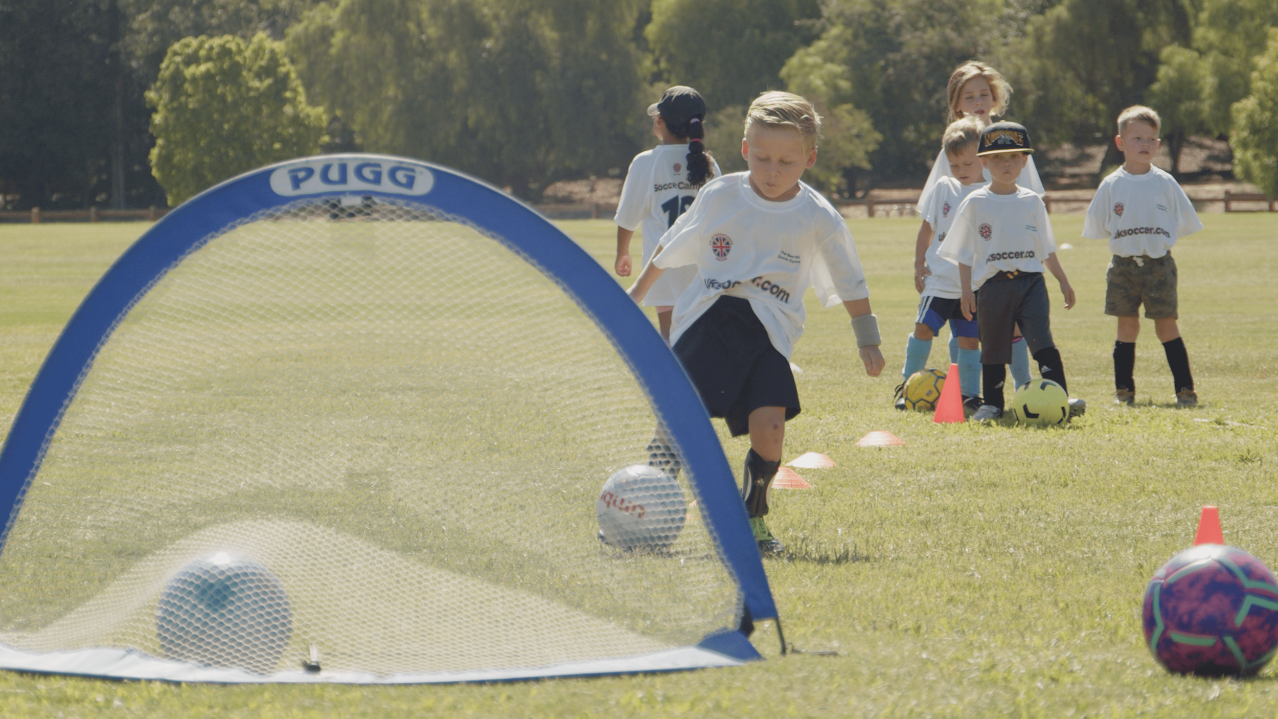 Young children participating in a soccer practice on a sunny day, with cones and a small goal on a grassy field.