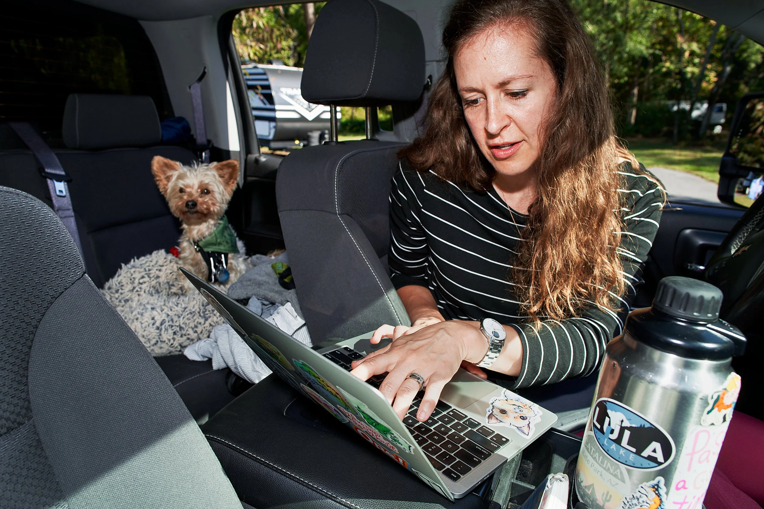 Remote worker (white woman with blonde, curly hair) using a laptop inside a parked vehicle with her small dog companion (short, blonde hair) in the back seat