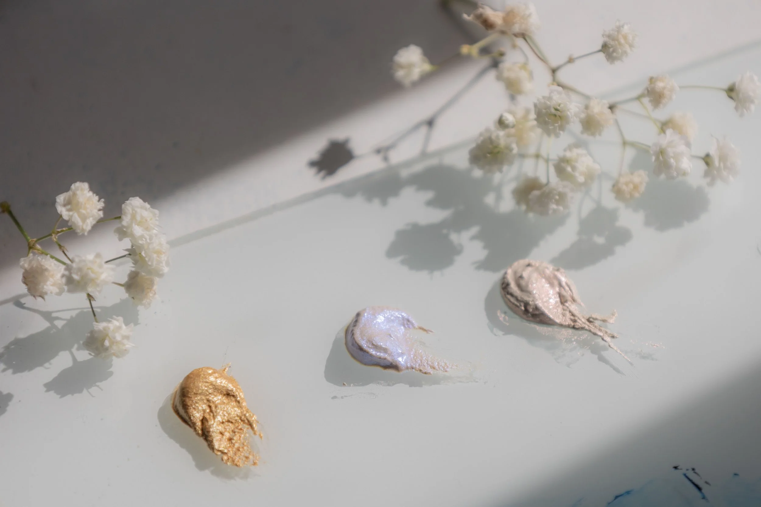 Several blobs of oil metallic paint in gold, silver, and bronze colors, along with some dried white flowers, are arranged on a white surface. Shadows cast by the flowers and paint blobs are visible. A palette from Love + Cross Studio.