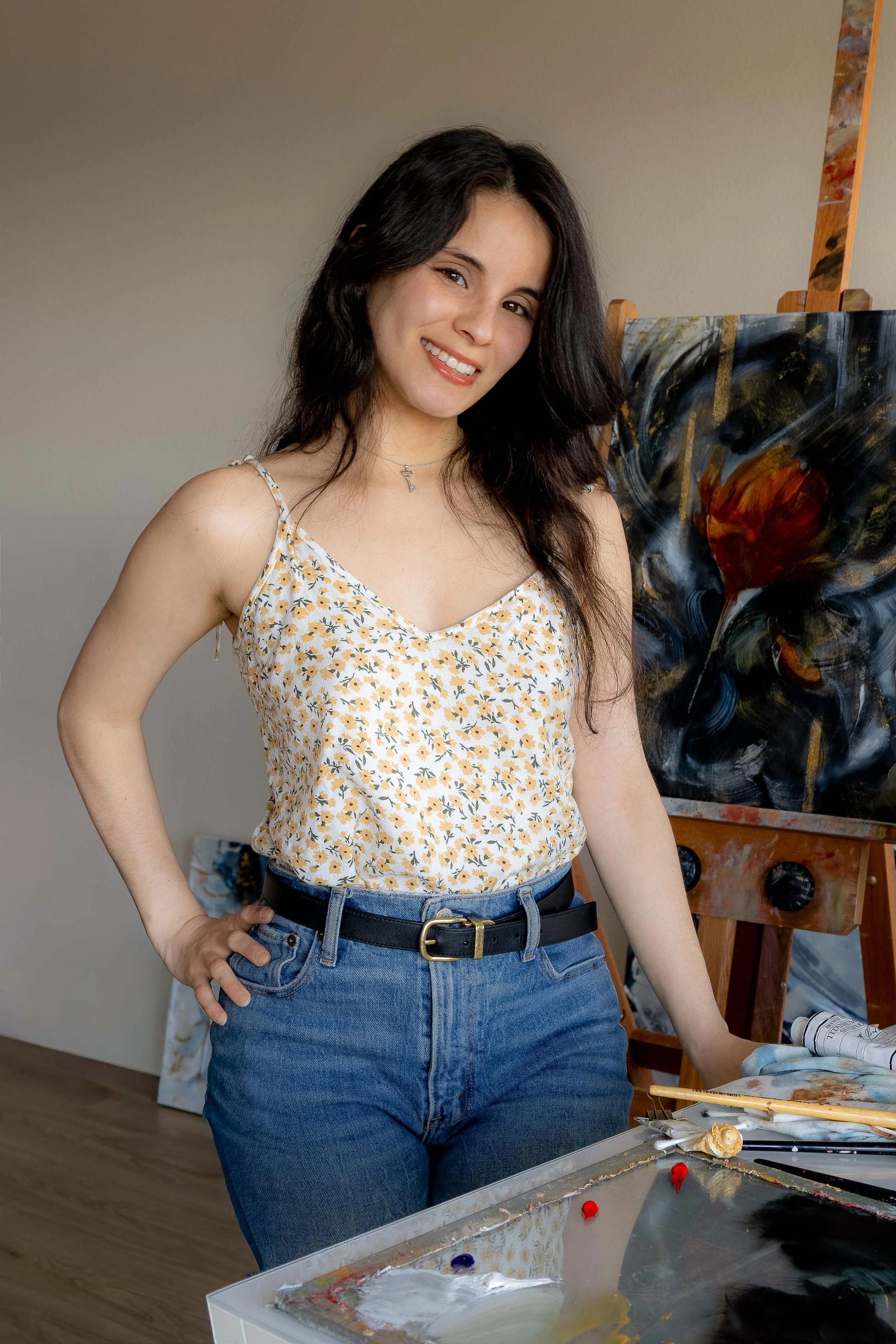 Artist Eliana of Love + Cross Studio  with dark hair, wearing a floral top and jeans, standing in front of an abstract painting on an easel in an art studio.