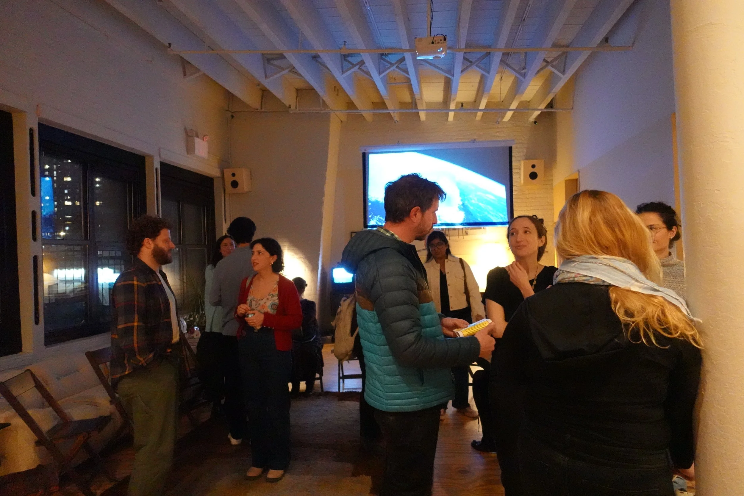 Event attendees talking near a large screen displaying a video, string lights overhead.