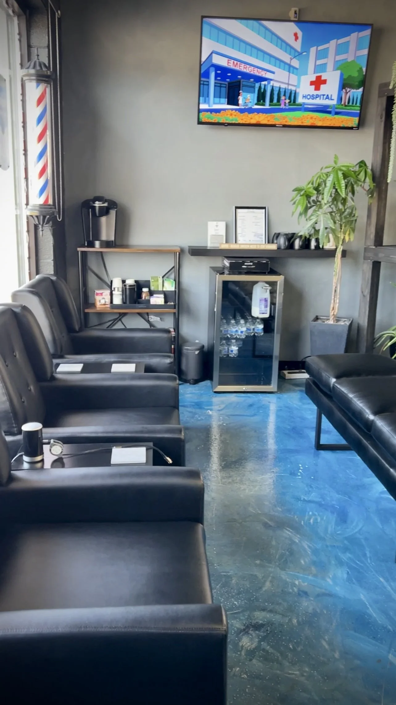 A modern waiting room with black leather chairs, a small water cooler, a mini fridge with bottled water, a potted plant, and a wall-mounted TV showing a hospital exterior. There is a barbershop pole decoration on the left.