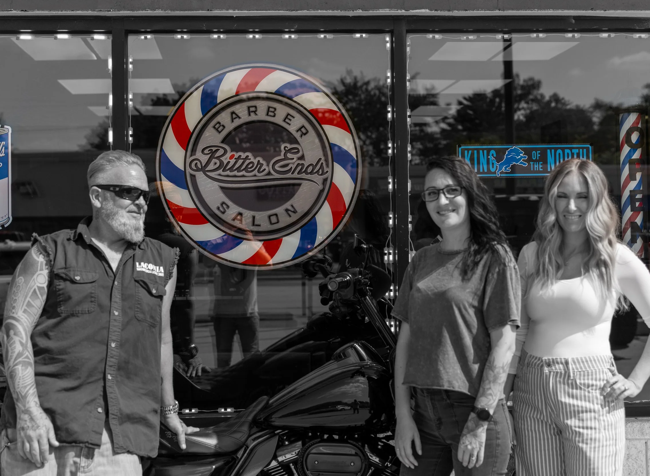 Three women and one man standing outside a barber shop called 'Bitter Ends' with a motorcycle in front of the window. The photo is mostly in black and white, with some color accents.