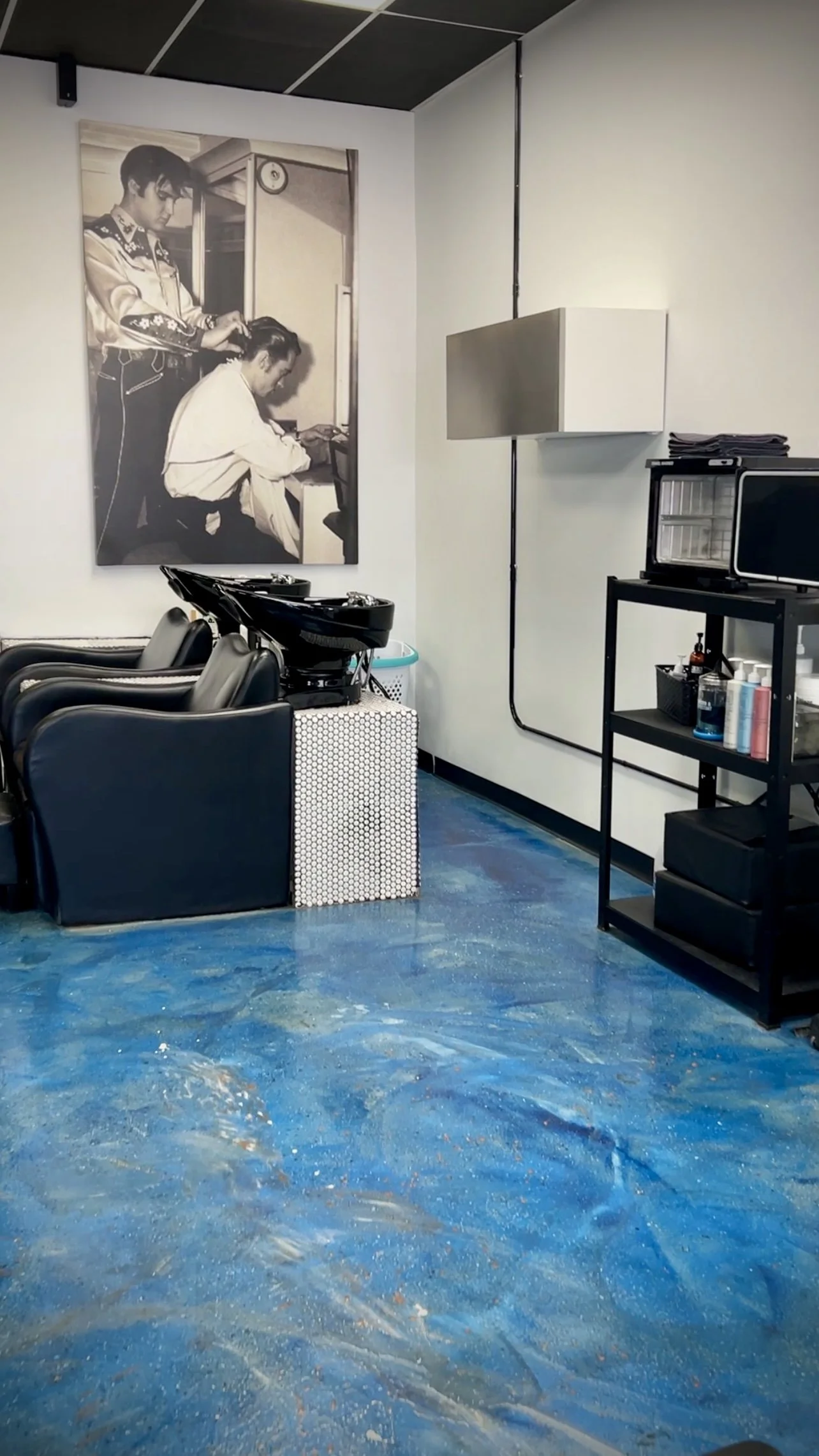 Salon station with black chairs, a print of a man getting his hair cut on the wall, and hair care products on a black cart.