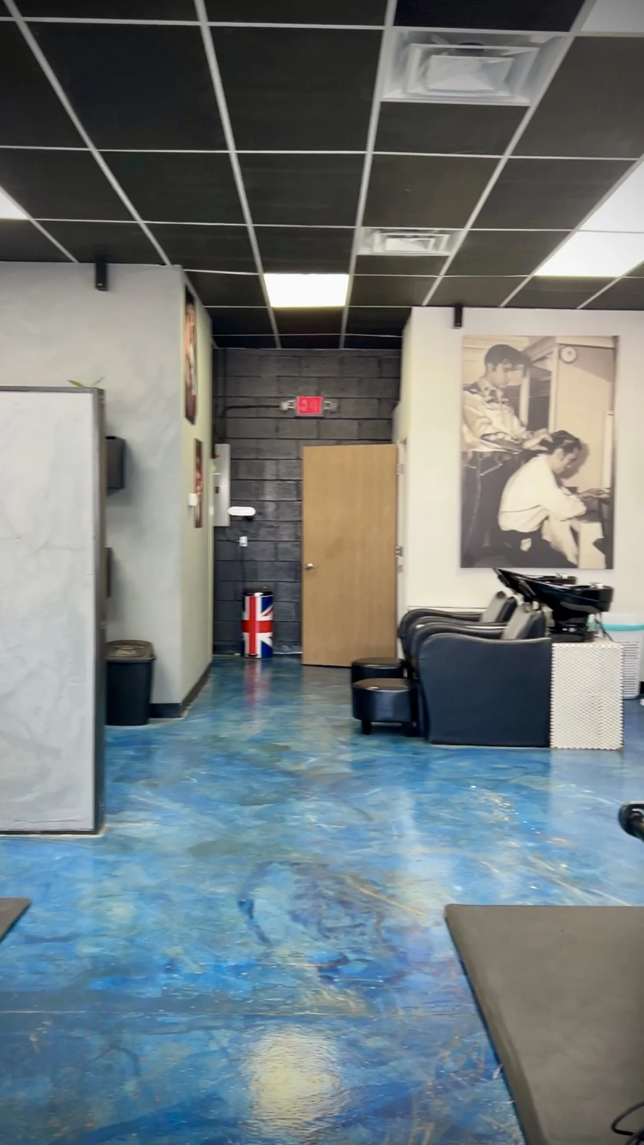 Inside a barbershop with black chairs and a hair washing station, a large black-and-white photo of children reading on the wall, a Union Jack trash can, and a digital clock on the wall.