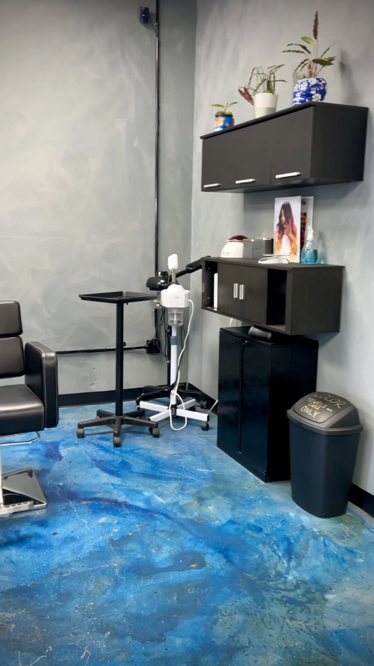 A salon or barber shop with black chairs, a rolling tray, and hair styling equipment. The wall is gray and has black shelves with decorative plants. The floor is painted with a blue, abstract design, and a trash can is visible.