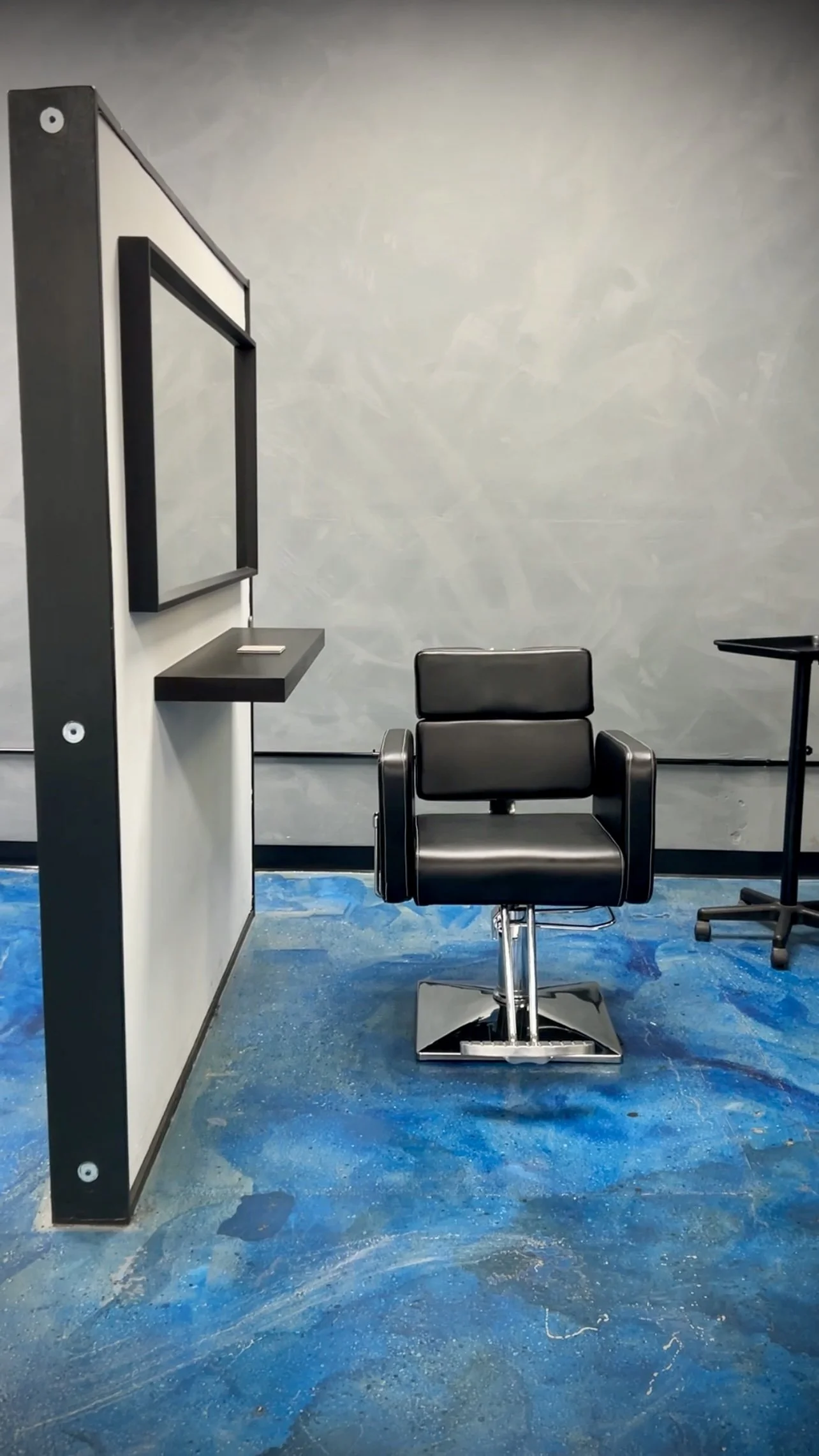 A black salon chair with a back and armrests in front of a plain gray wall, on a blue marbled floor, with a mirror and small shelf on a partition to the left and a small black table to the right.