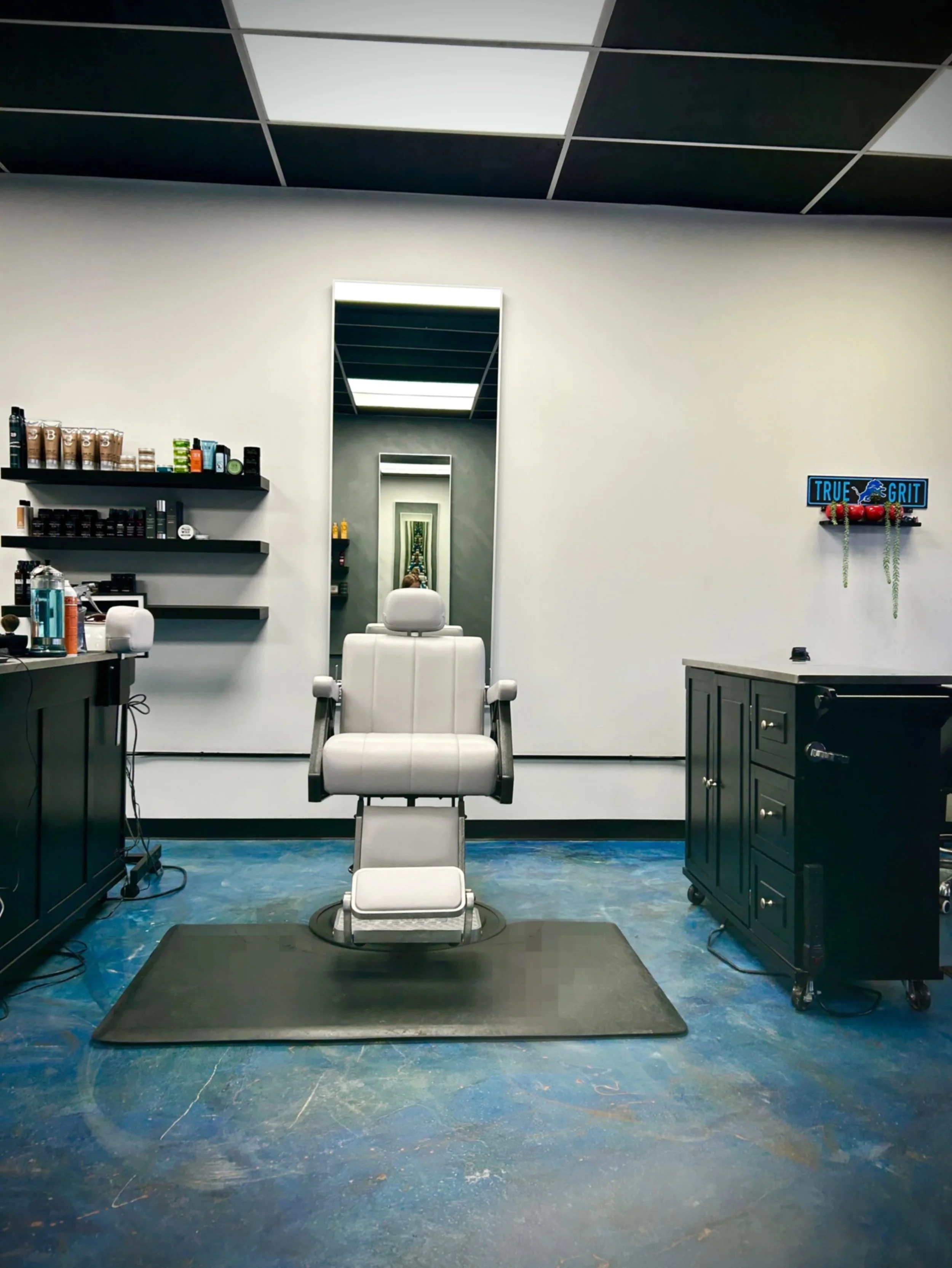 Empty barber's or salon chair in front of a mirror in a minimalistic salon or barber shop setting with shelves of hair products on the left wall and a 'True Grit' sign on the right wall.