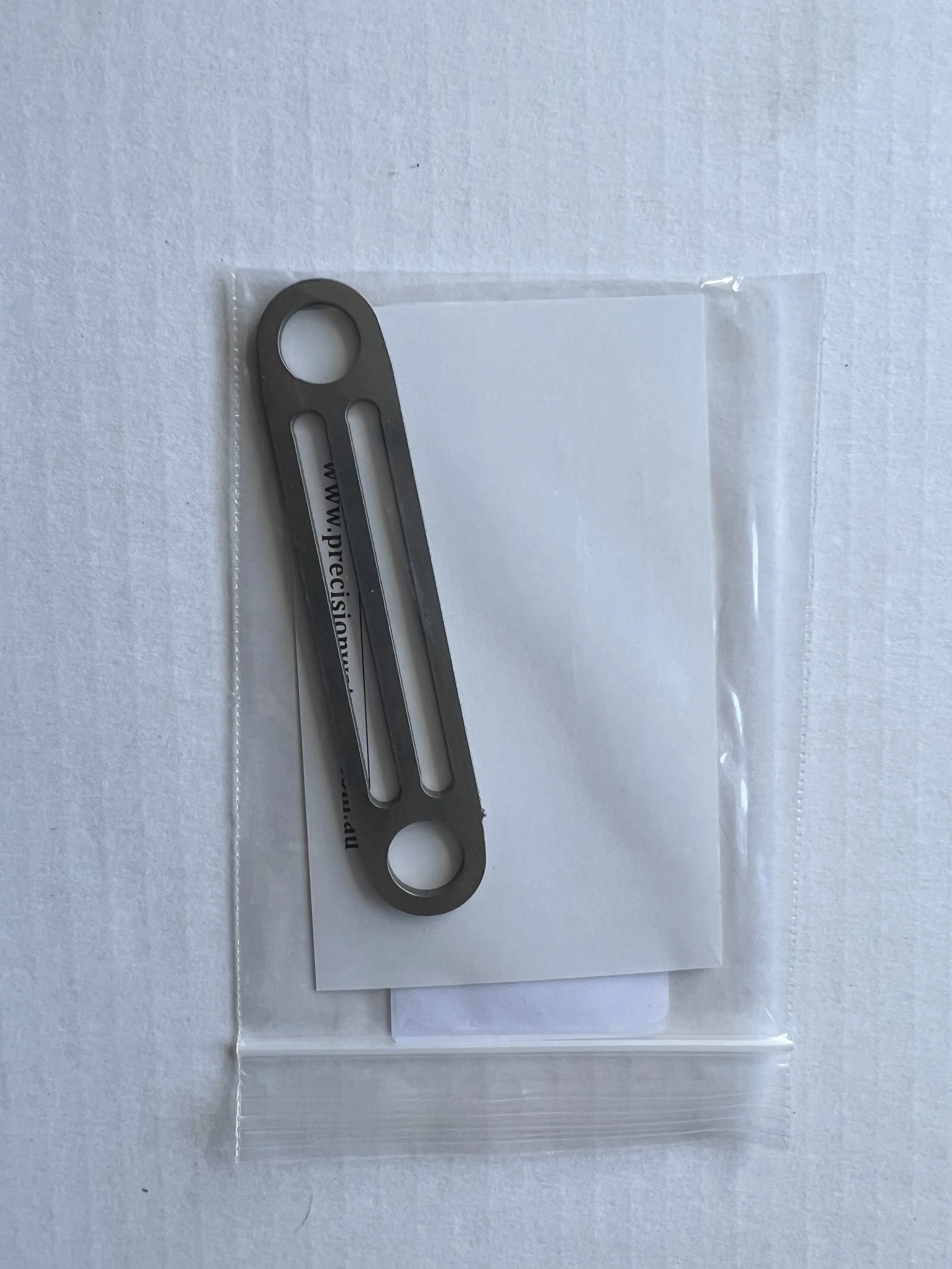Small metallic tool with two circular holes and two elongated slots, labeled with a website URL, placed on a piece of white paper inside a plastic bag.