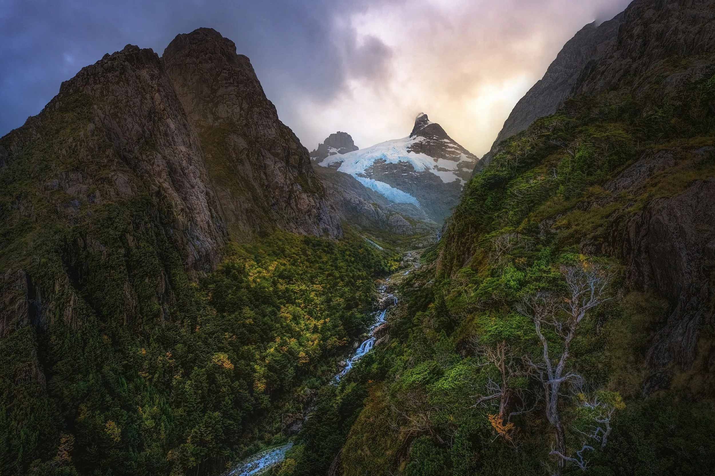 Majestic mountain valley with snow-capped peaks, dense green forest, and a waterfall flowing through the valley, under a cloudy sky with hints of sunlight.