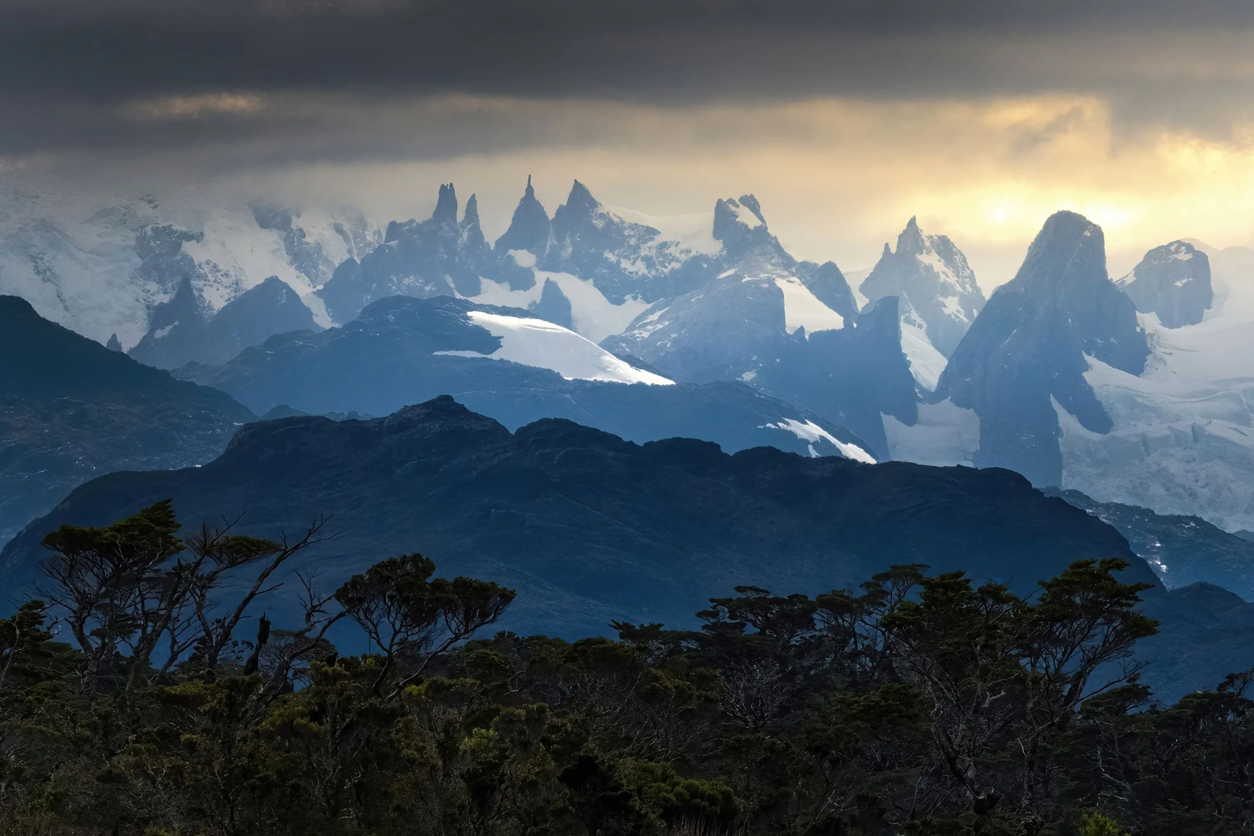 Mountain range with snow-capped peaks and dark clouds, trees in the foreground.