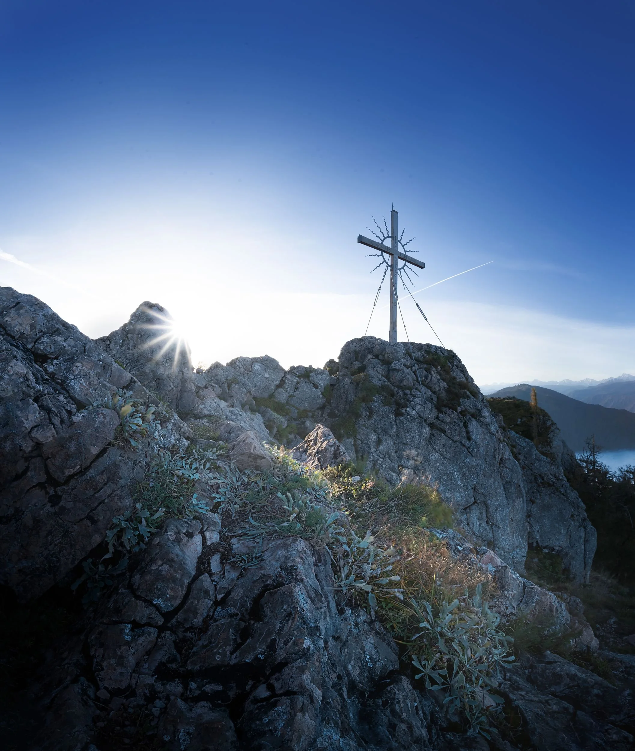 Rugged mountain peak with a metal cross at the summit, sunlight shining behind rocks and sparse vegetation, with a clear blue sky and distant mountain range in the background.
