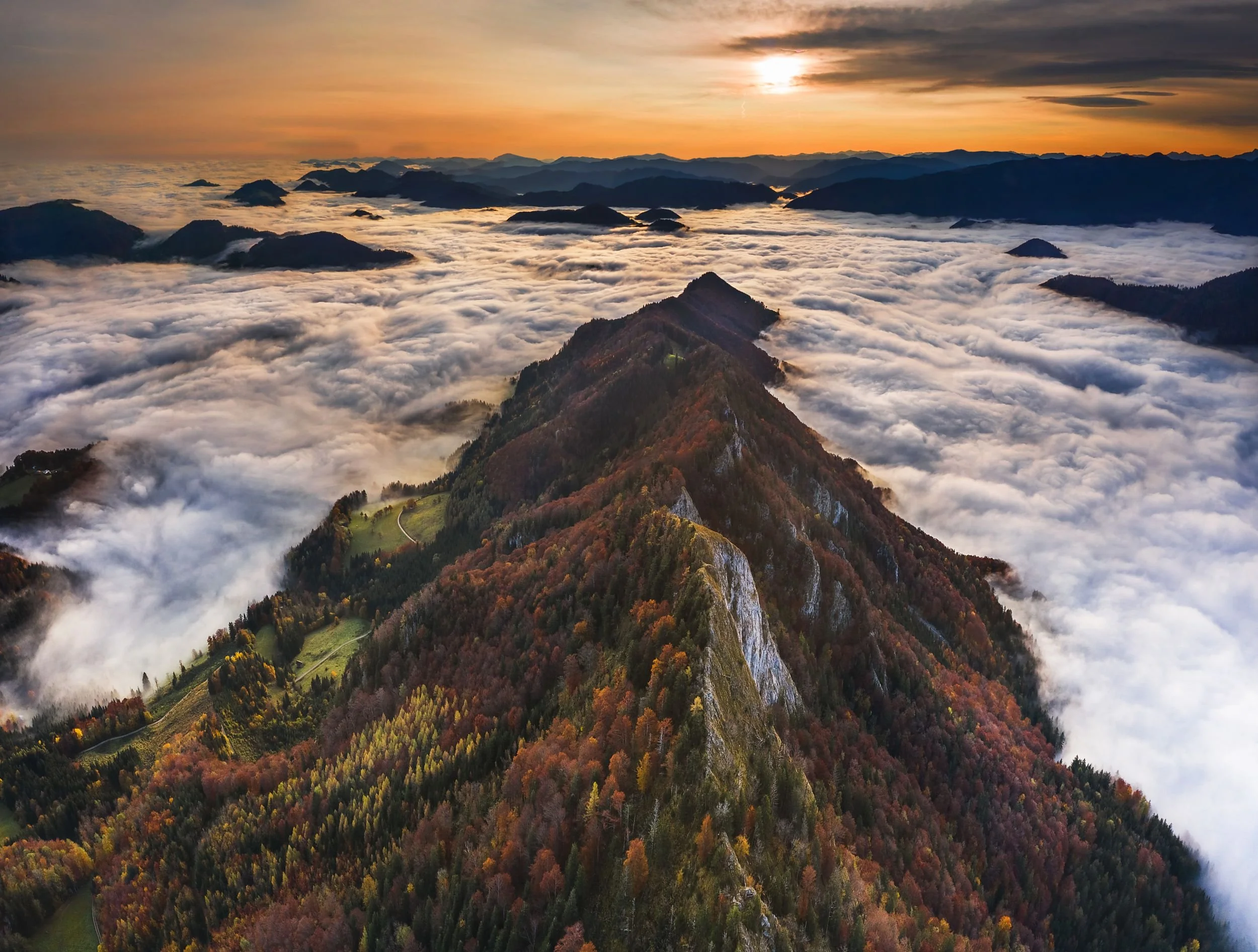 Aerial view of a mountain ridge covered with colorful trees and a green valley below, with clouds surrounding the peaks and a sunset sky in the background.