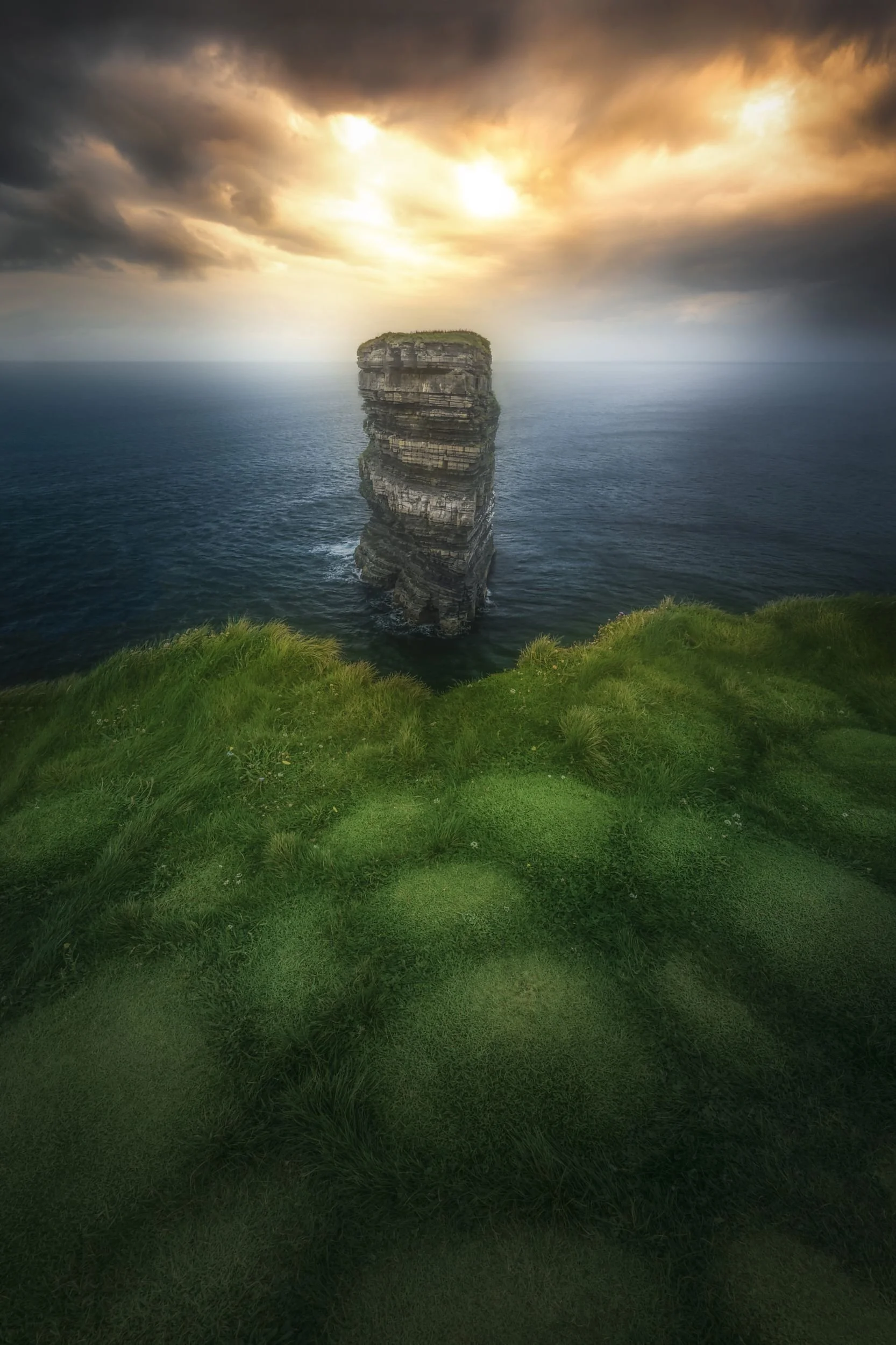 Cliffside view of a tall, layered sea stack in the ocean, with green grass in the foreground and a dramatic cloudy sky at sunset.