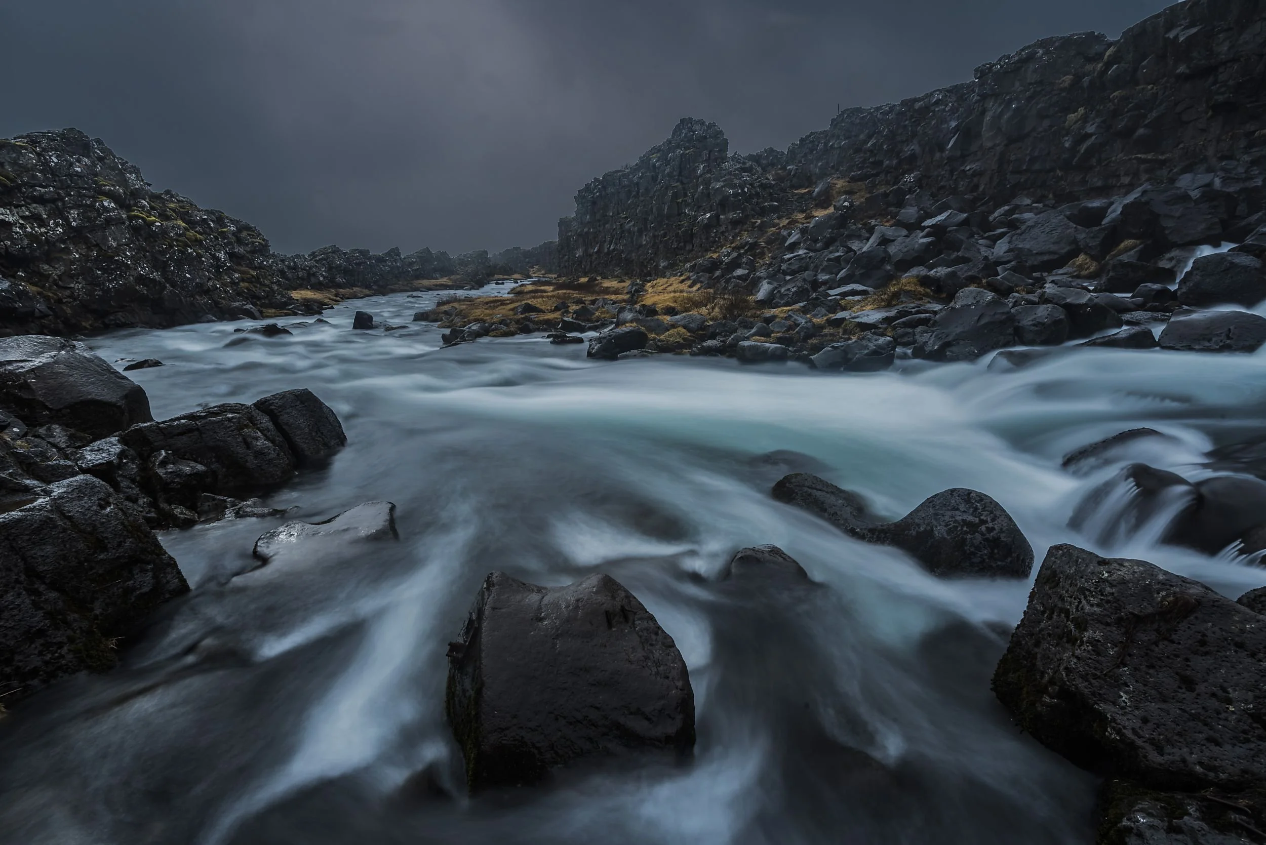Raging river running through a rocky canyon with dark cliffs and cloudy sky