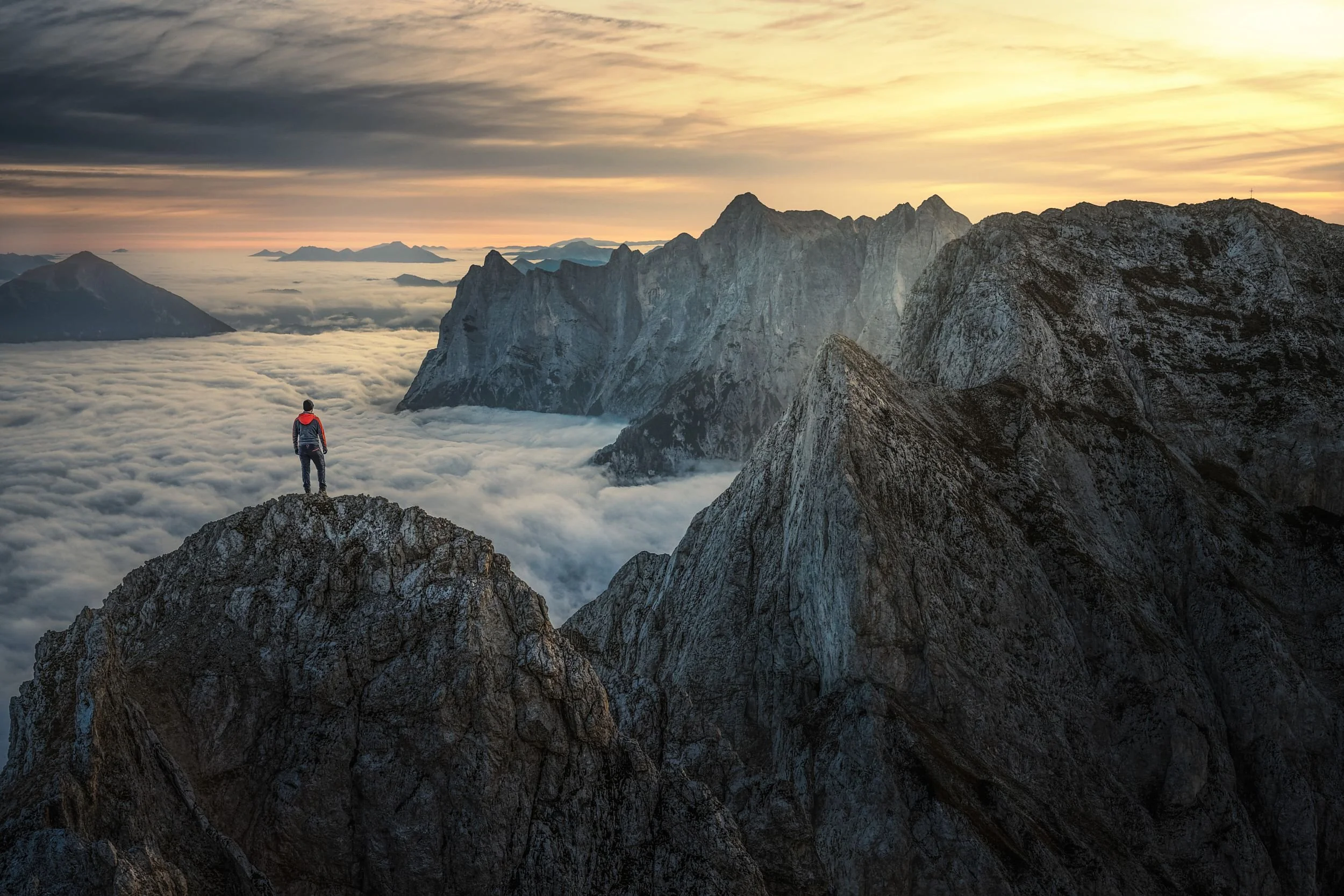 A person stands alone on a rocky mountain peak overlooking a sea of clouds and rugged mountain ridges under a colorful, cloudy sky at sunset or sunrise.