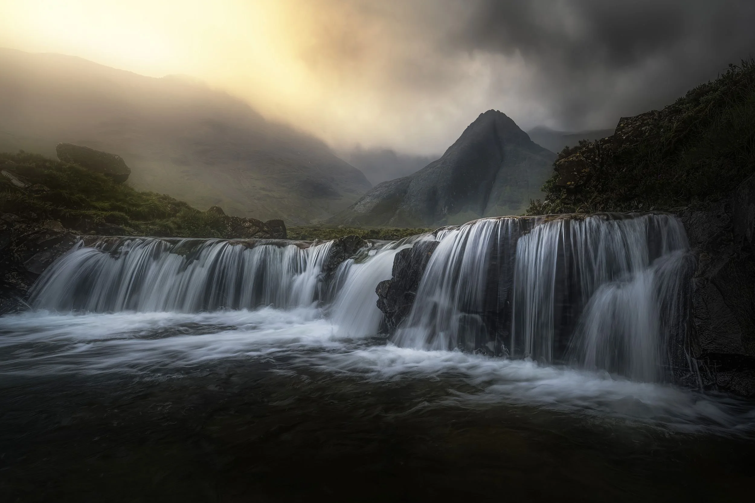 A mountain landscape with a waterfall in the foreground, misty mountains in the background, and a cloudy sky.