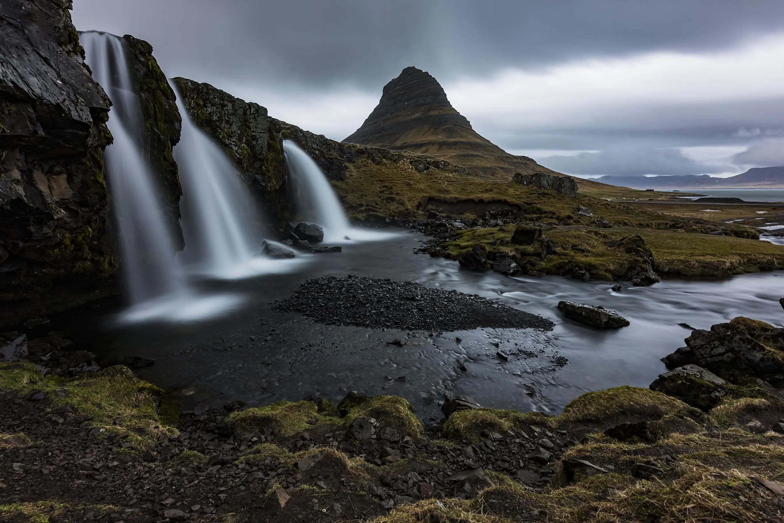 A landscape featuring a waterfall cascading over rocks, with a mountain in the background and moss-covered ground.