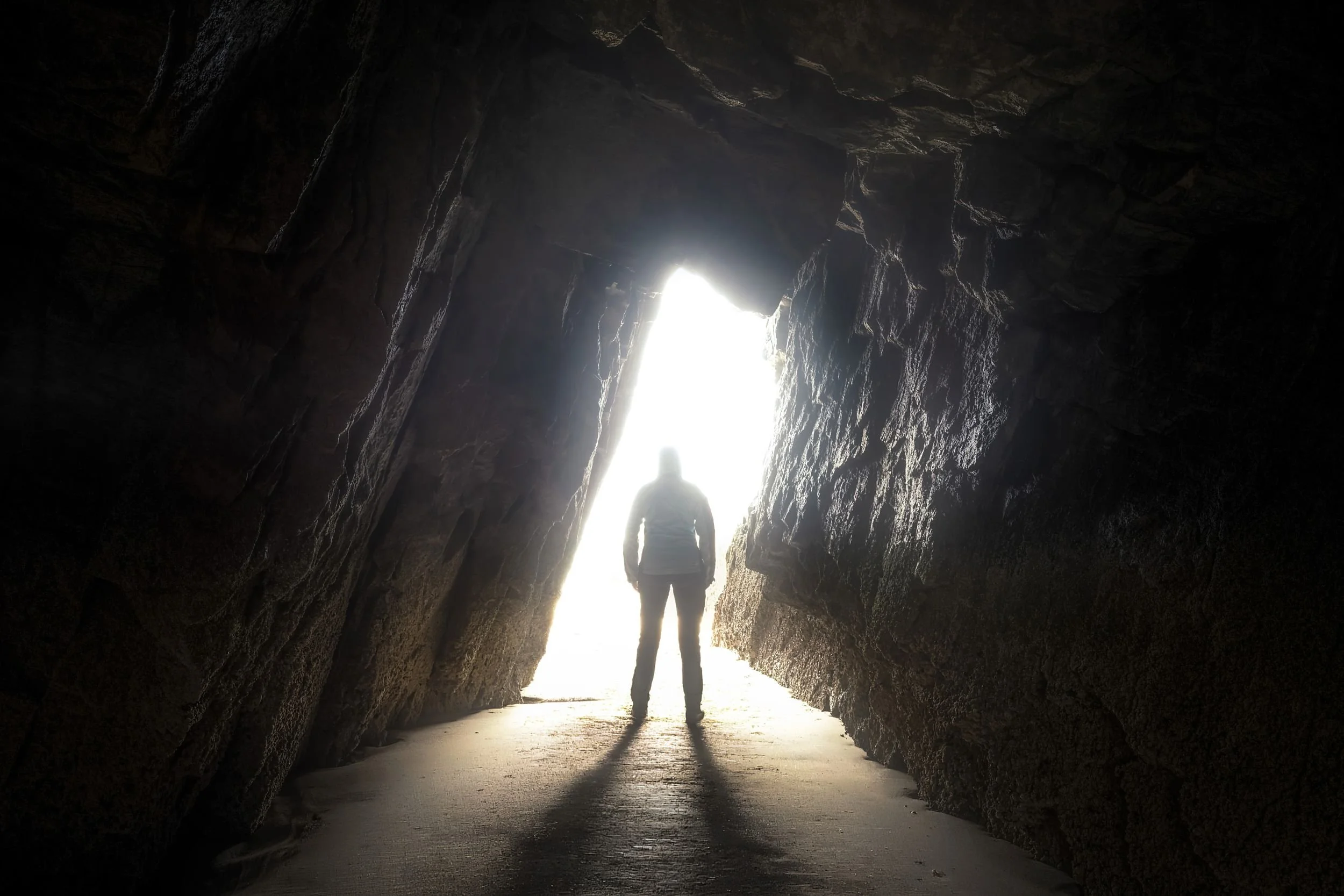 Eine Person, die in einer dunklen Höhle steht, blickt hinaus auf den hell erleuchteten Eingang, durch den Tageslicht strömt.