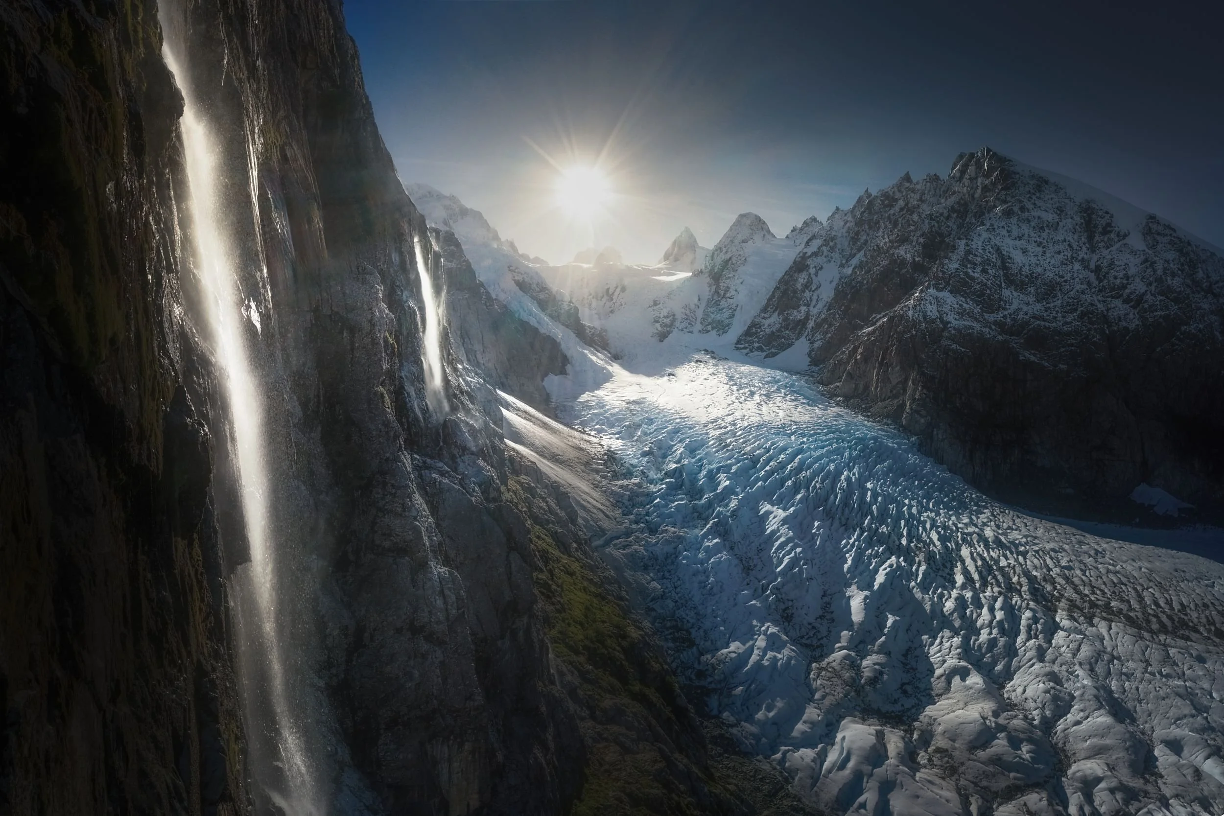 Snow-capped mountains with glaciers, a waterfall cascading down a rocky cliff, and the sun shining over the landscape.