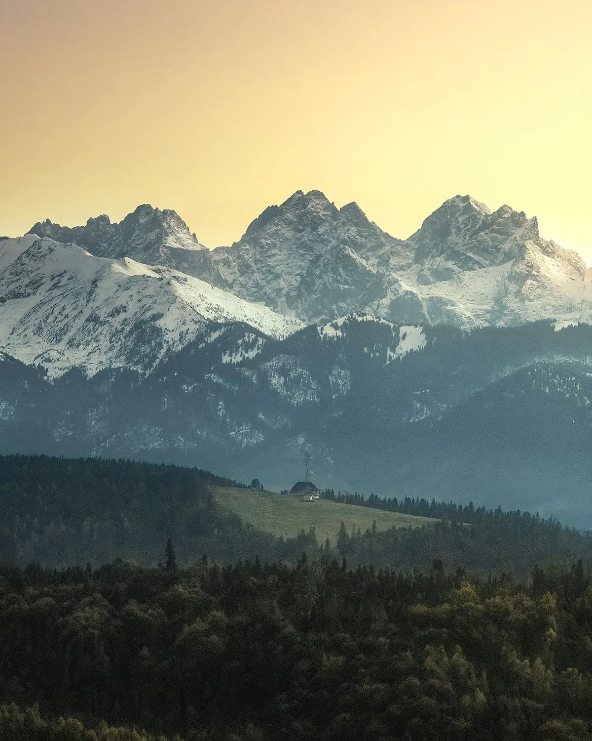 Snow-capped mountain range with several peaks under a yellow sky, forested hills, and a small house or hut in the foreground.