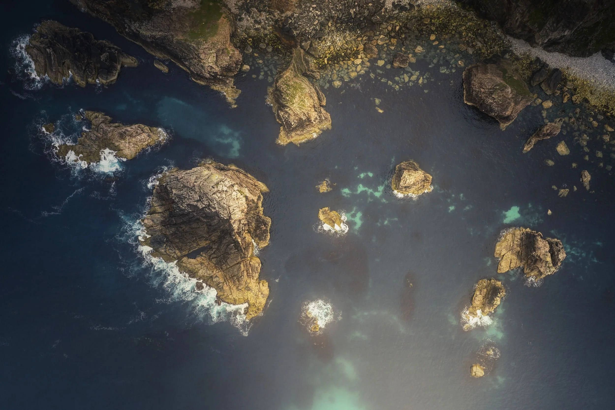 Aerial view of large rocks and small islands in the ocean near a rocky shoreline with some green moss or algae.