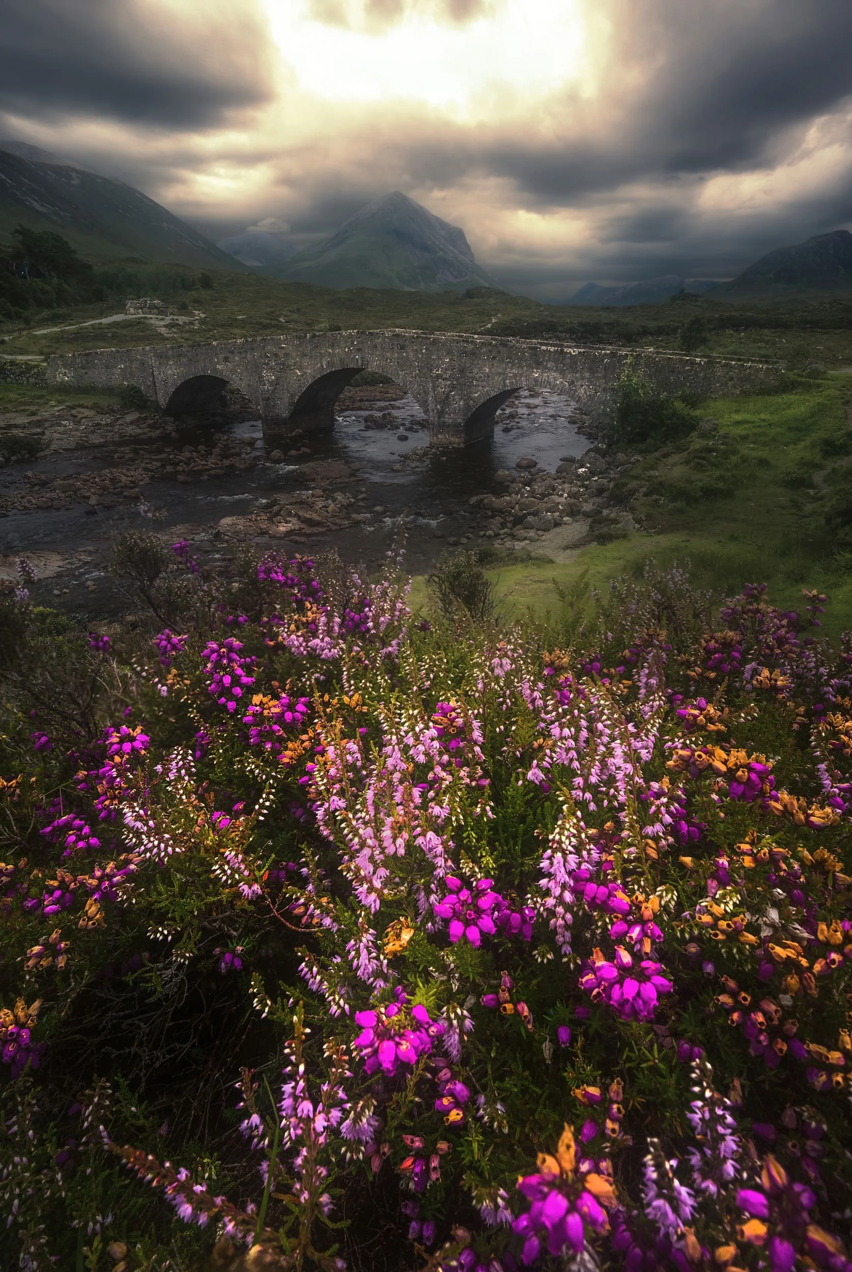 Bunte Blumen im Vordergrund, ein Steinbogenbrücke über einen Fluss, Berge und Wolken im Hintergrund bei bewölktem Himmel.