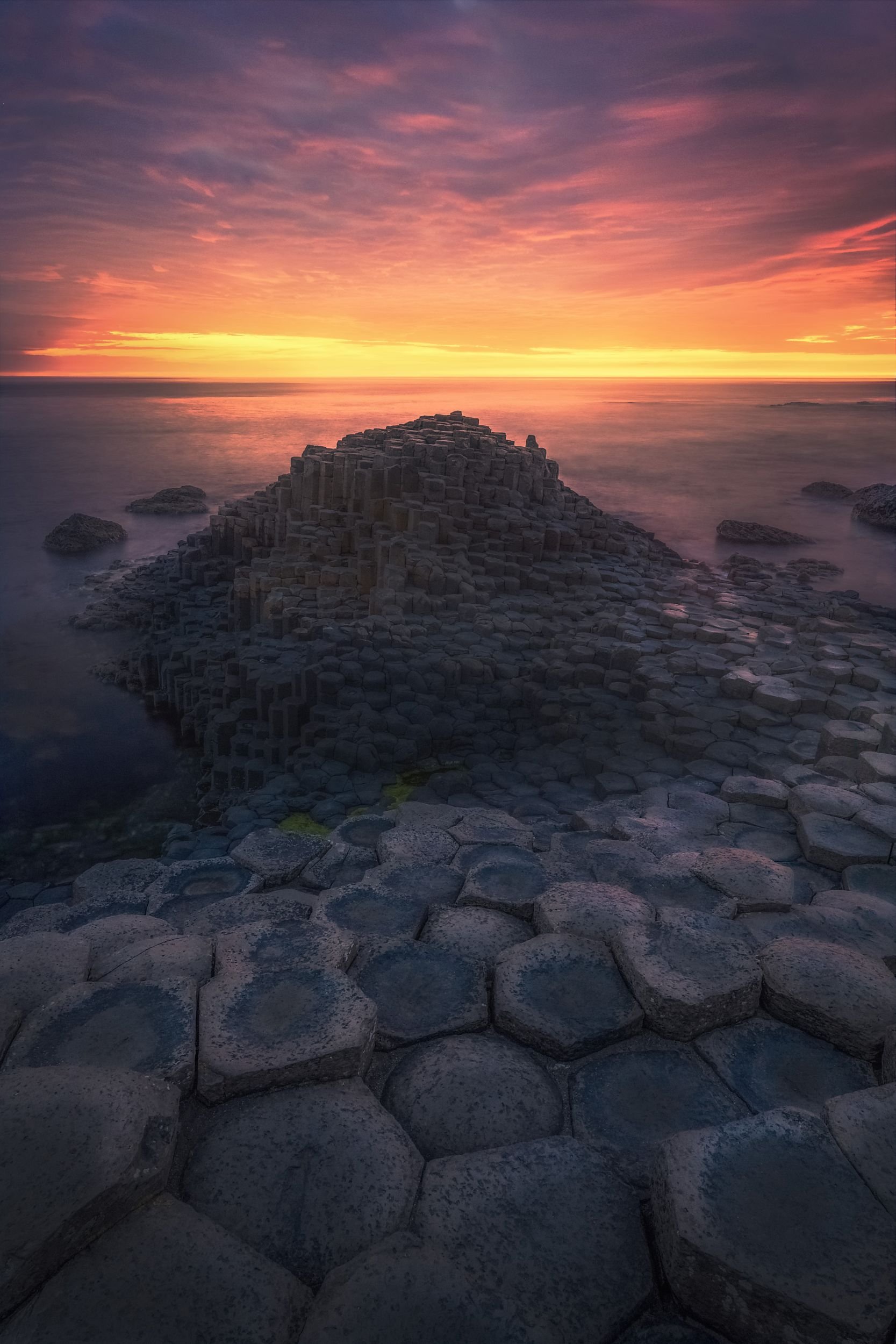 Sunset over the ocean with stone formations in the foreground, including a large pyramid-shaped structure made of hexagonal basalt columns.