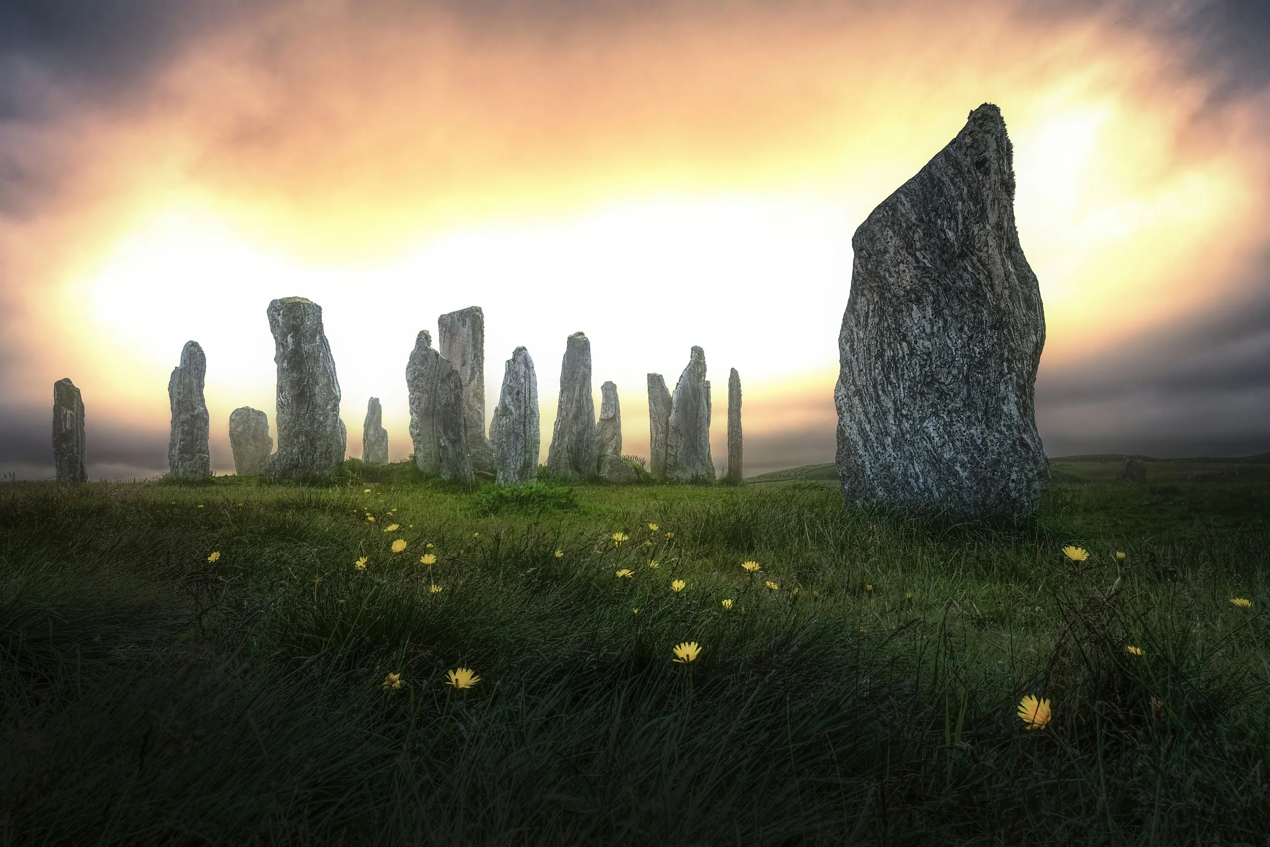 A row of ancient standing stones on a grassy hill with yellow flowers, under a sunset sky with dark clouds.