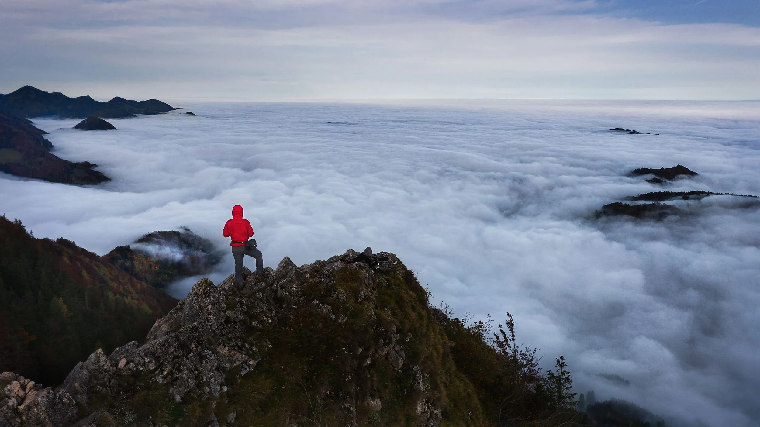 Person in a red jacket standing on rocky mountain peak overlooking a sea of clouds and distant mountains.