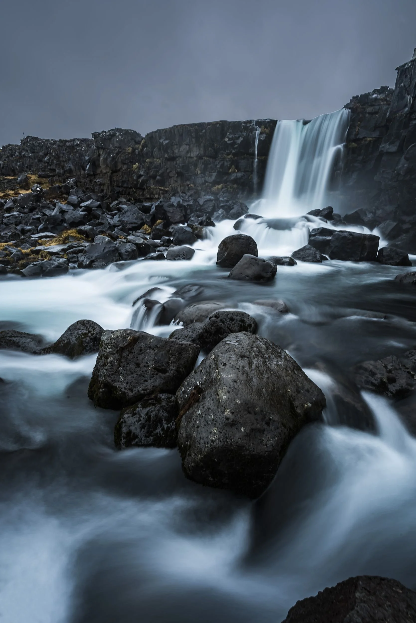 A waterfall flowing over dark rocks in a rugged, misty mountain landscape.