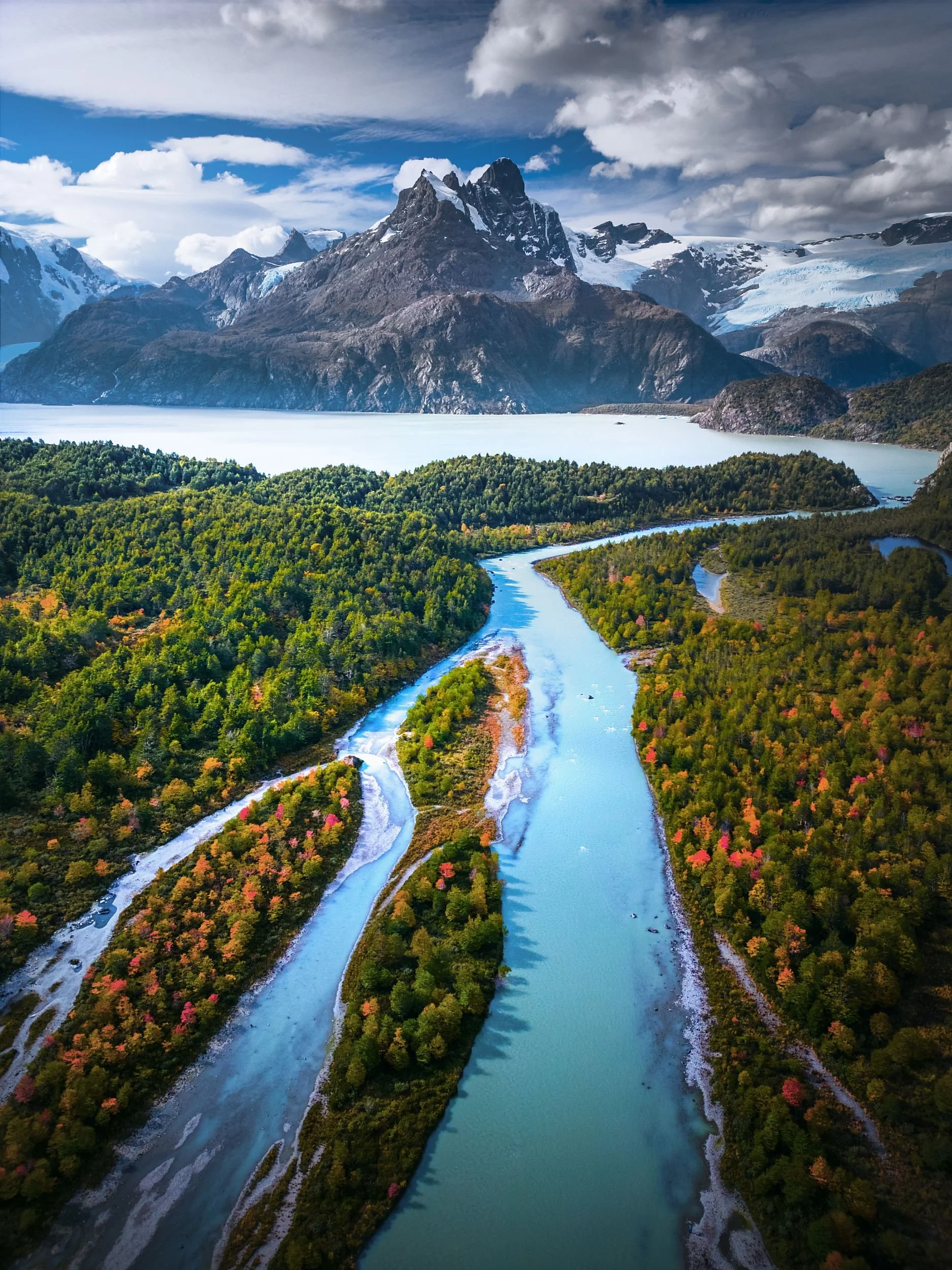 Aerial view of a river winding through a lush green forest with snow-capped mountains and glaciers in the background under a partly cloudy sky.