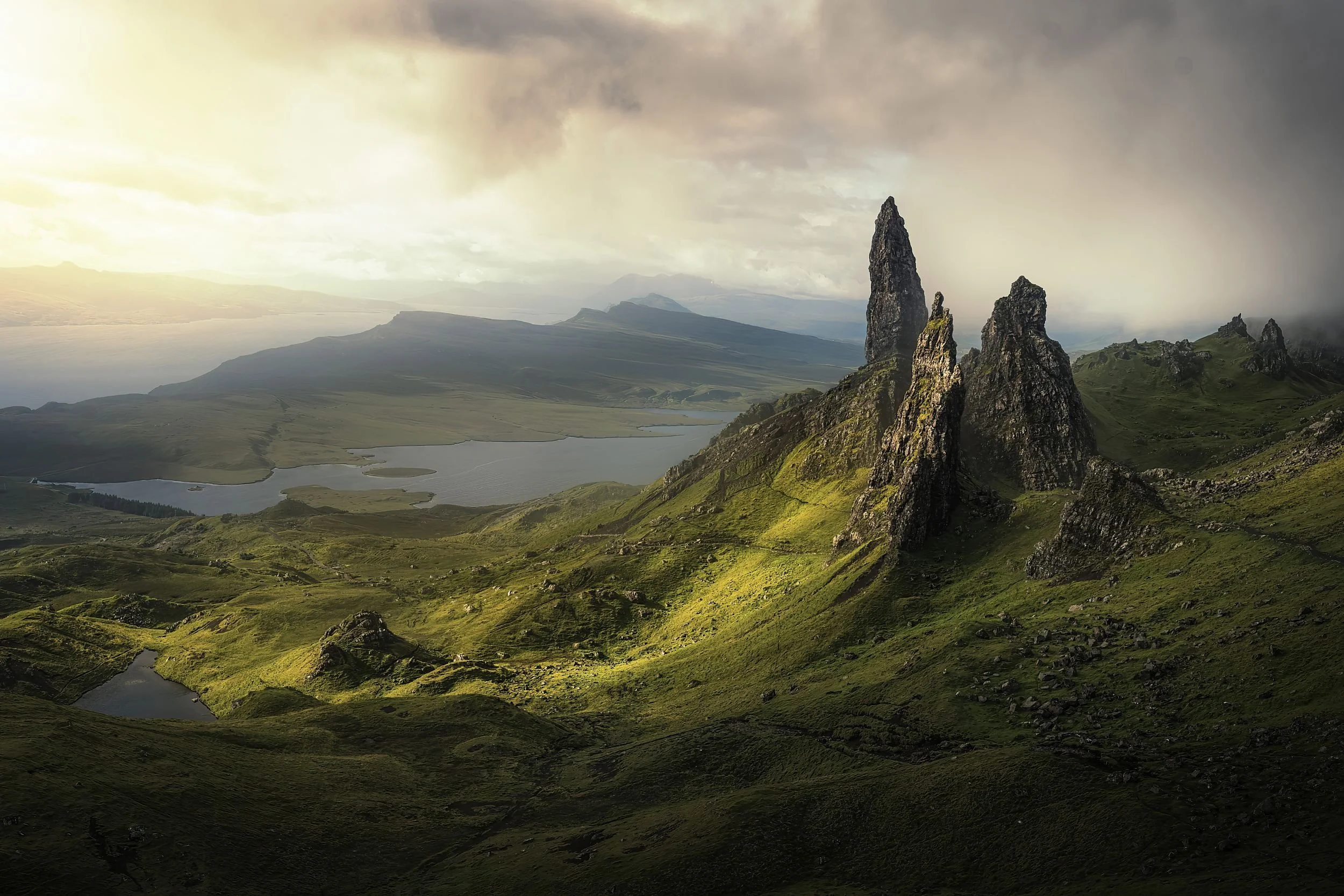 Scenic landscape of rugged green hills and towering rocky formations with a lake in the background under a cloudy sky, soft sunlight illuminating parts of the terrain.