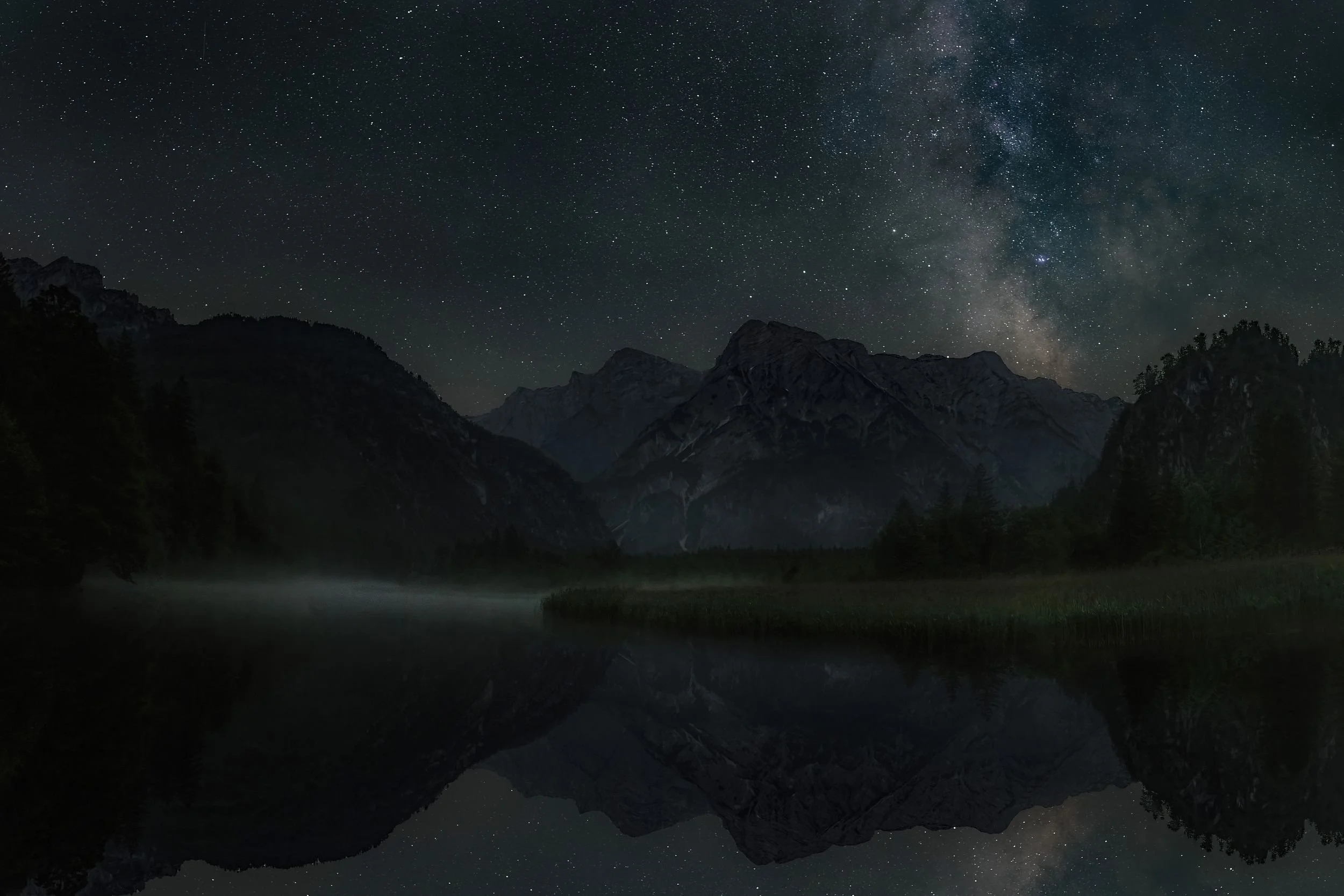 Nighttime landscape of mountains and forest reflected in a calm lake with a starry sky and the Milky Way galaxy overhead.